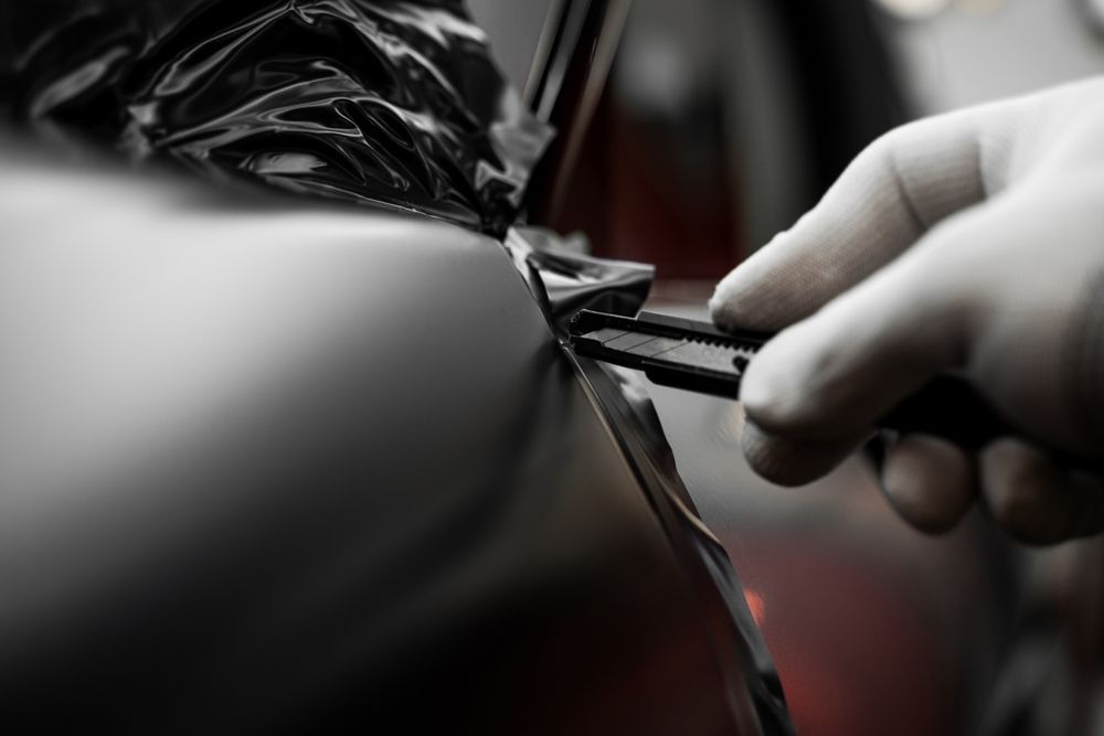 A gloved hand uses a utility knife to carefully trim black vinyl wrap along the edge of a vehicle's body panel.