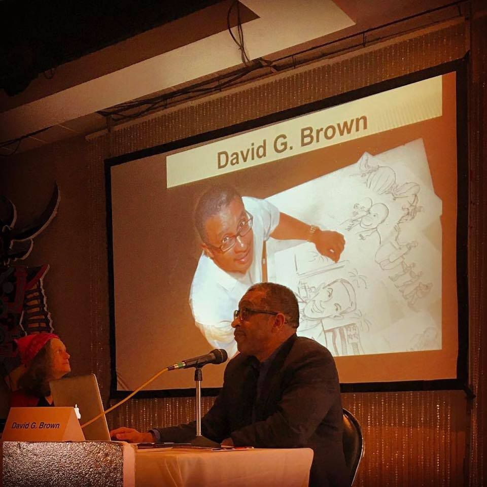 Man at a podium with a projector screen showing a photo of him drawing. Another person sits on the left.
