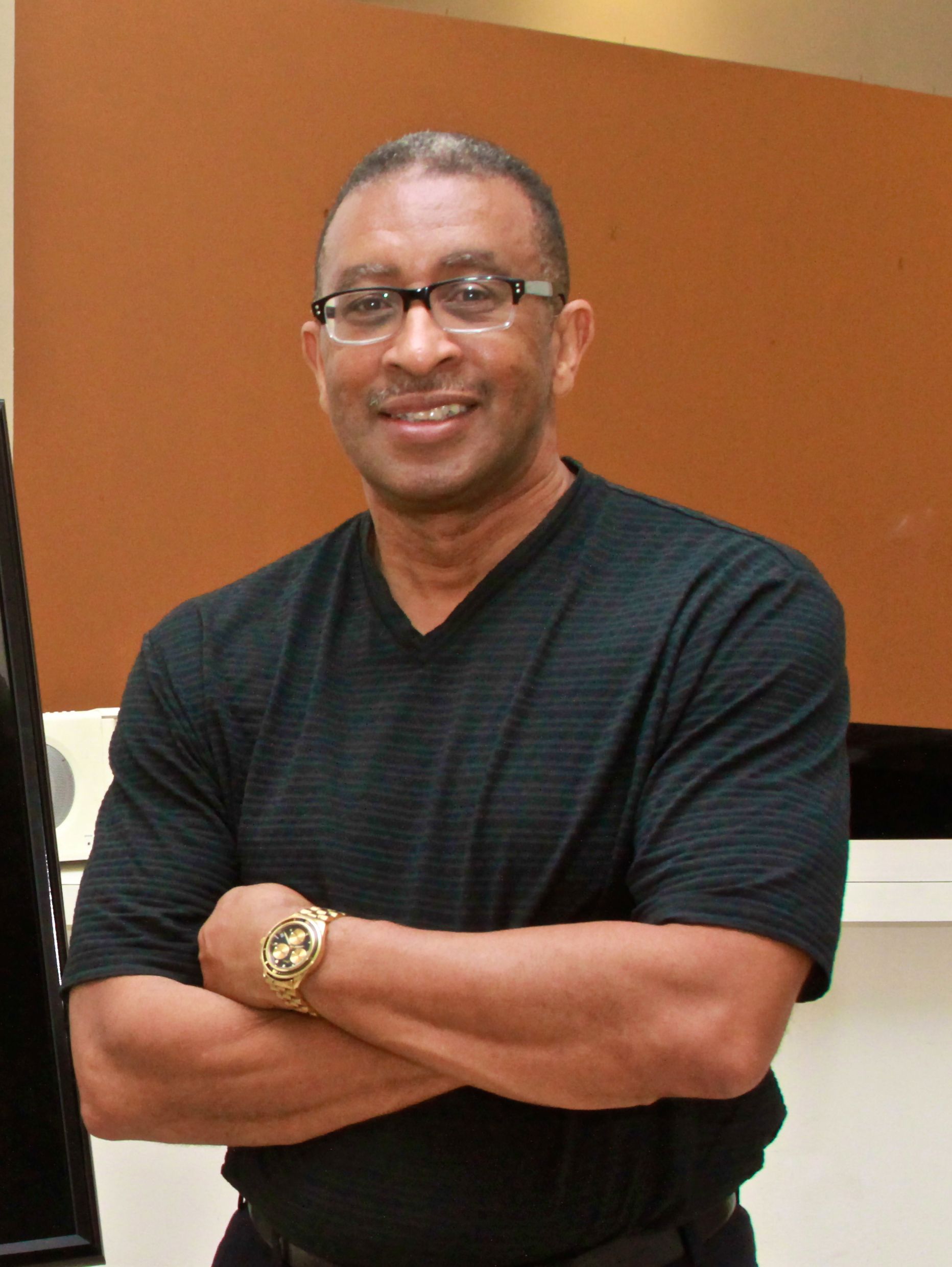 Man with glasses smiles, arms crossed, wearing a black shirt and a gold watch. Brown background.