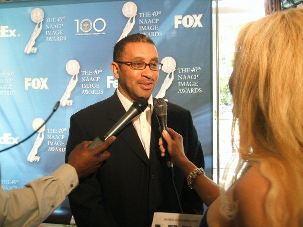 Man in suit being interviewed at the NAACP Image Awards. Microphones held by two people. Blue backdrop.