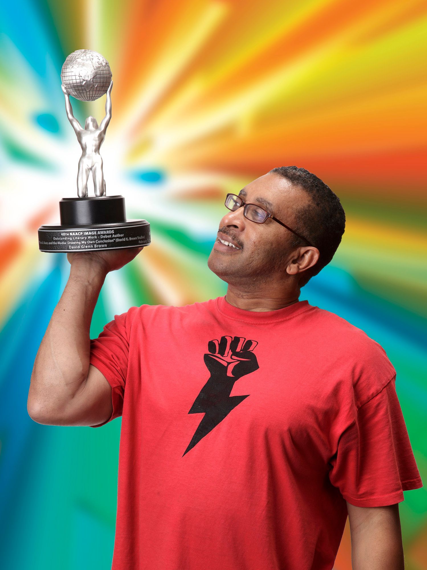 Man in red shirt holding award, looking up. Colorful background.