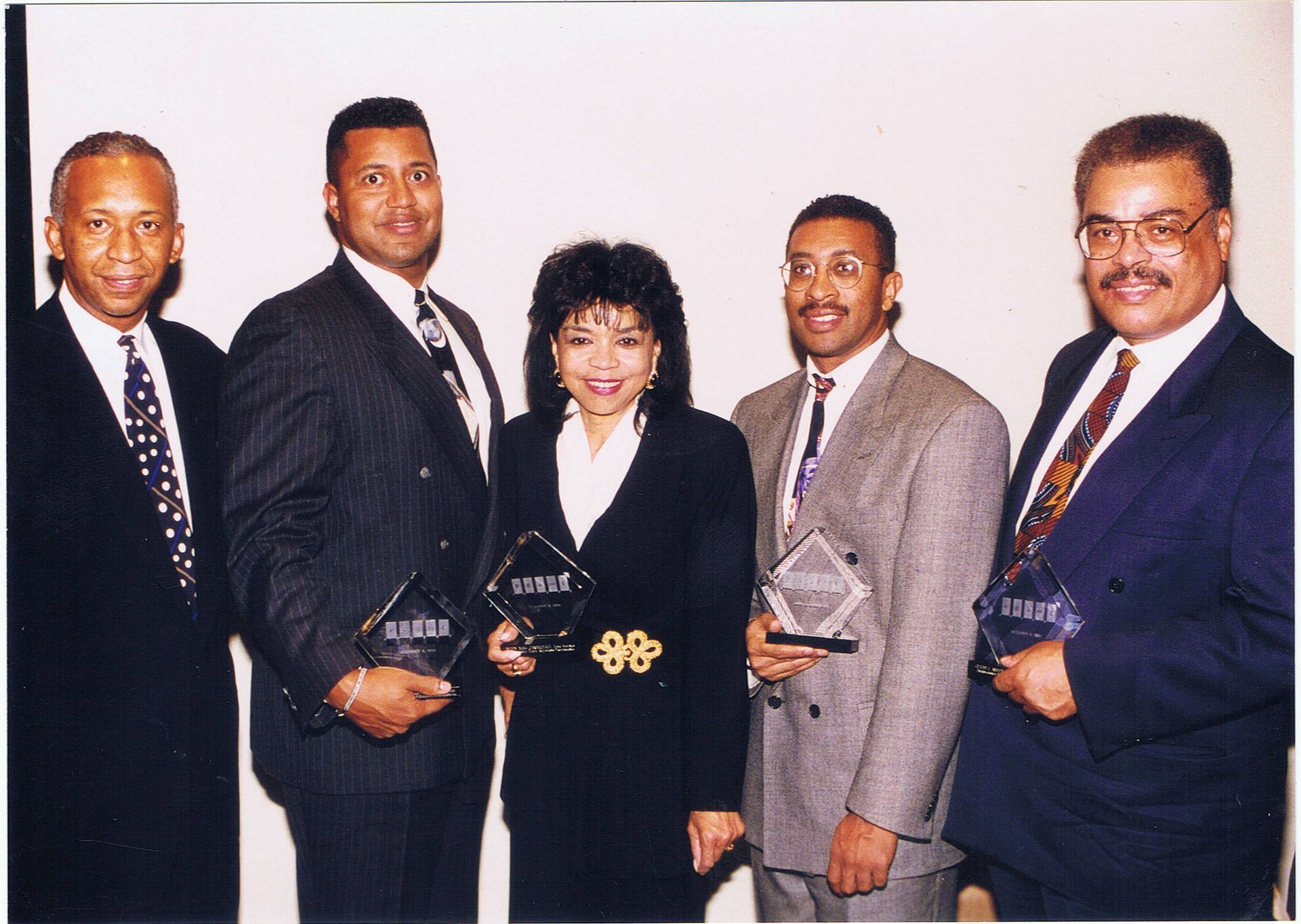 Five people in business suits holding awards, smiling, standing in front of a white wall.