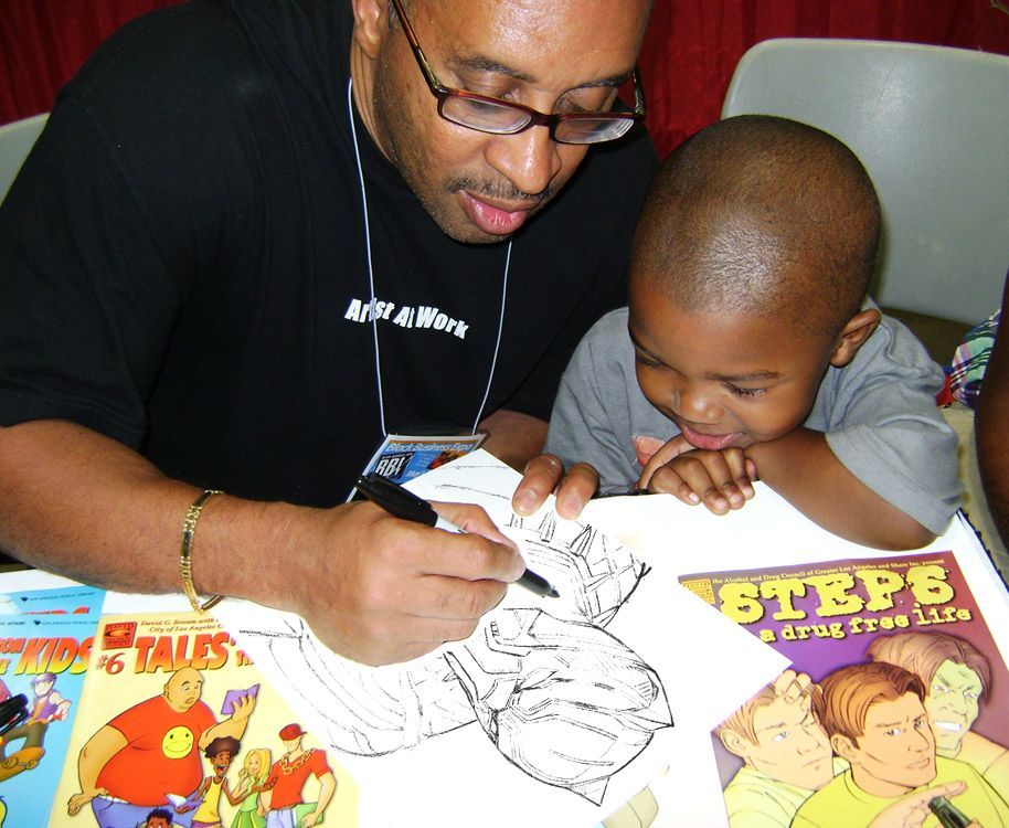 Man with glasses drawing in a comic book with a young boy, books in foreground, at an event.