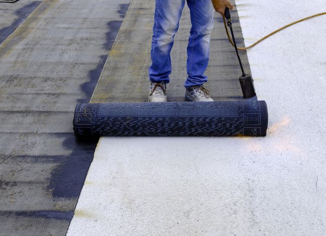 Trabajador aplicando una capa de revestimiento negro para techos sobre una superficie de techo blanca.