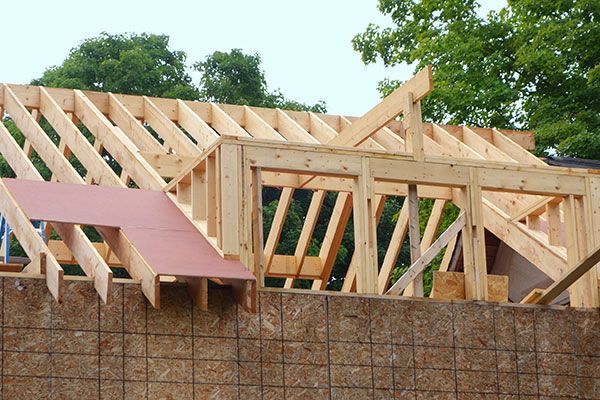 Wooden house frame under construction. Roof trusses and wall supports visible against a cloudy sky.