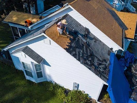 Workers on a roof removing old shingles. White house with a blue tarp.
