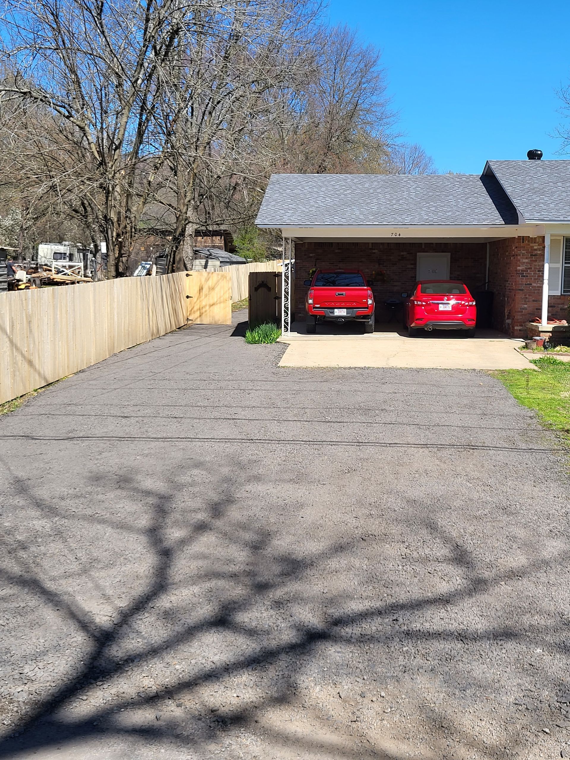 Gravel driveway with two red cars in a carport. A wooden fence lines the left. Bright sunlight, blue sky.
