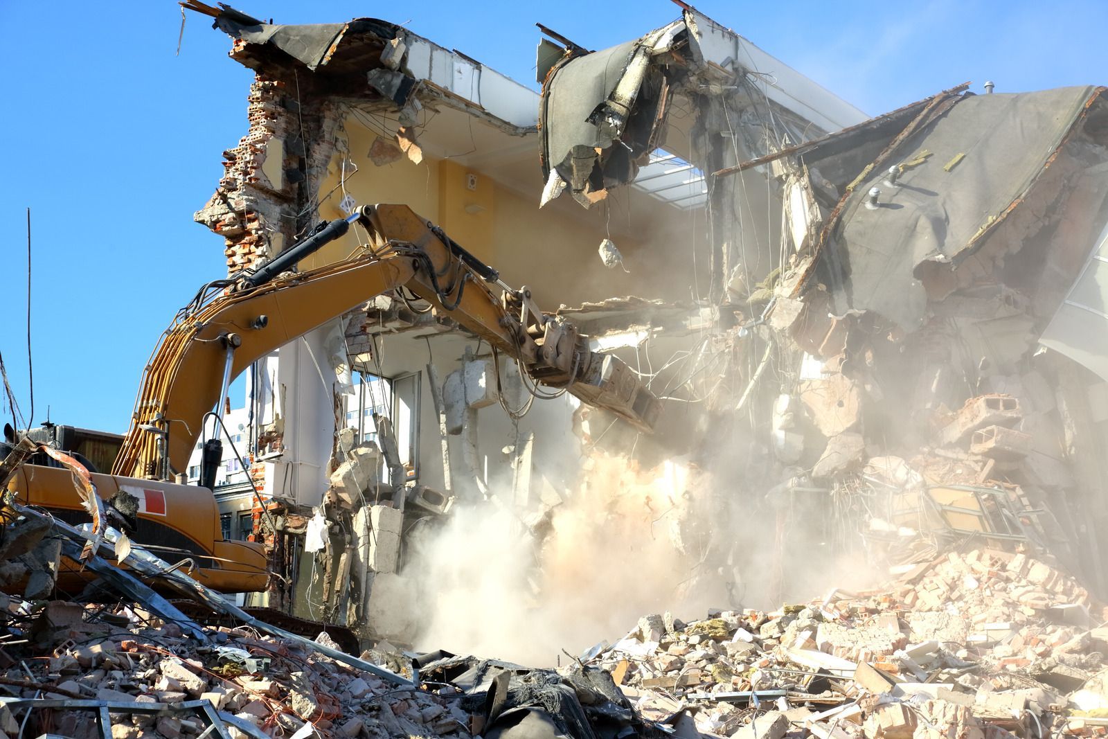 A building being demolished by an excavator, creating a dust cloud under a blue sky.