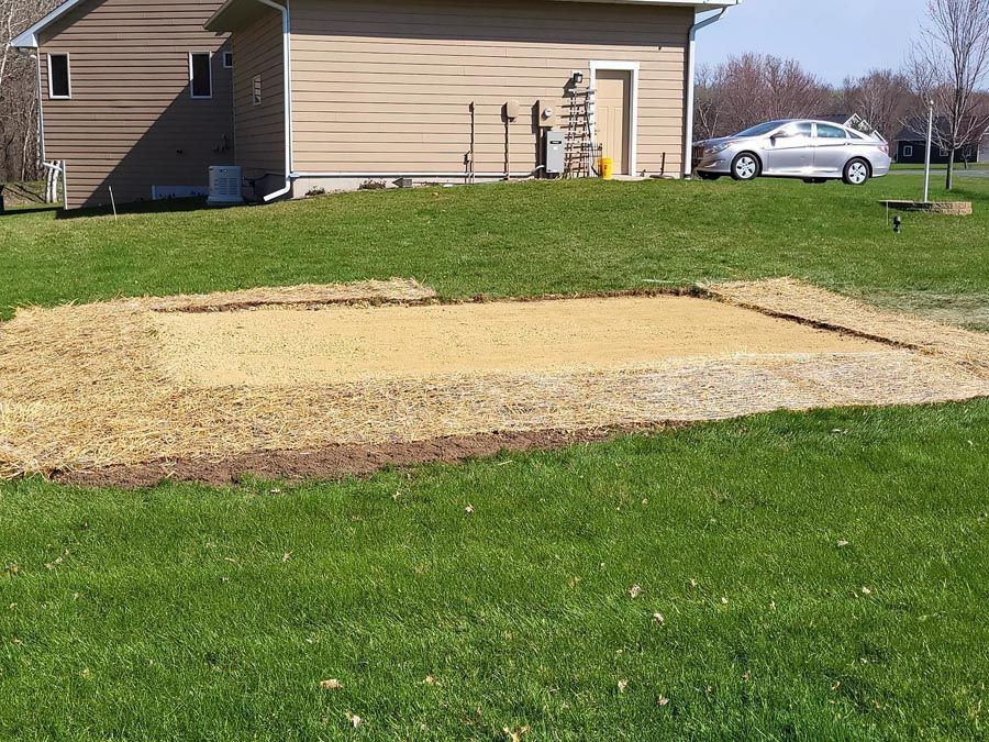 A rectangular area of gravel and dirt is in a grassy yard, next to a beige house and a car.