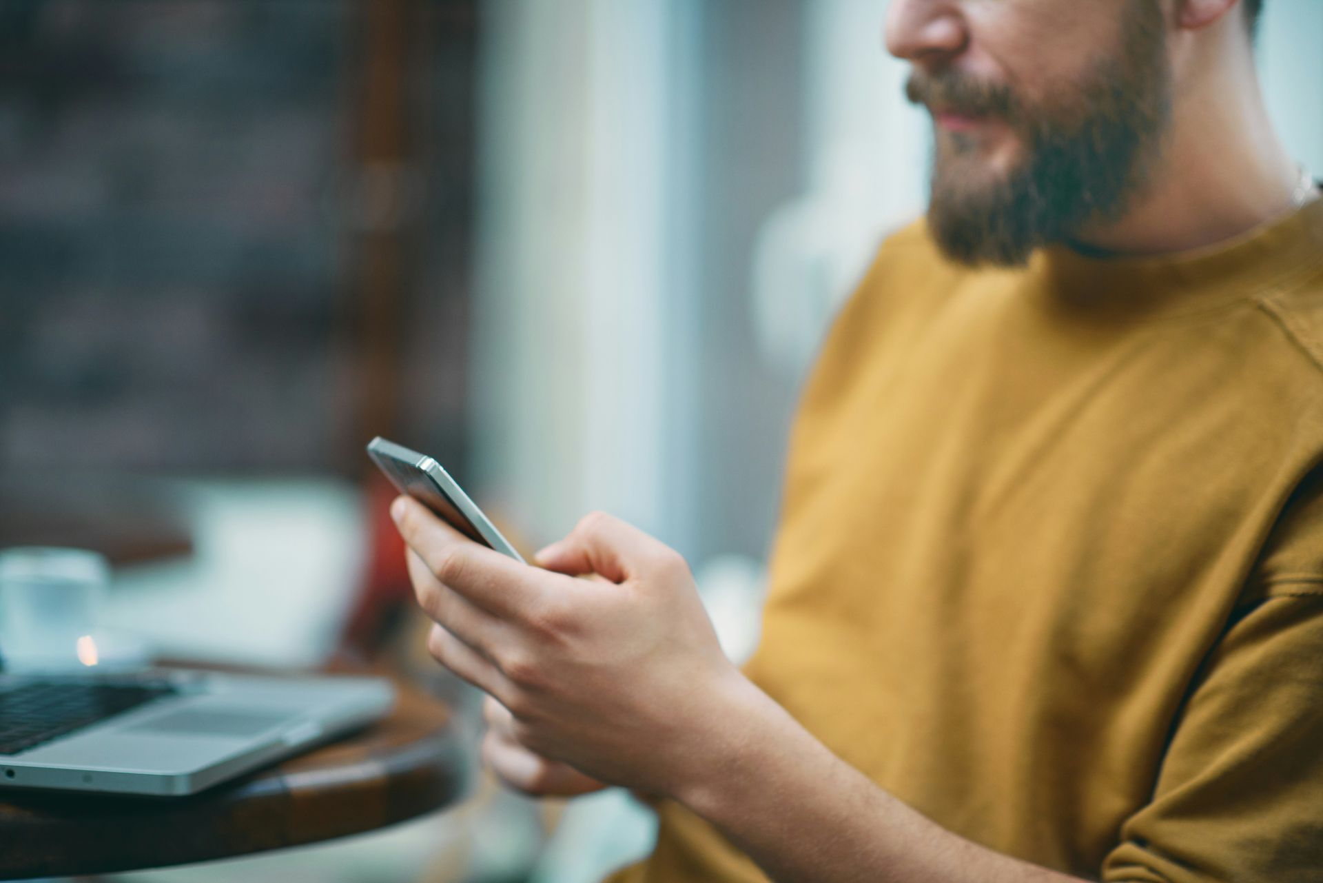 Man with beard using a smartphone; he wears a yellow sweater and sits near a laptop in a cafe.