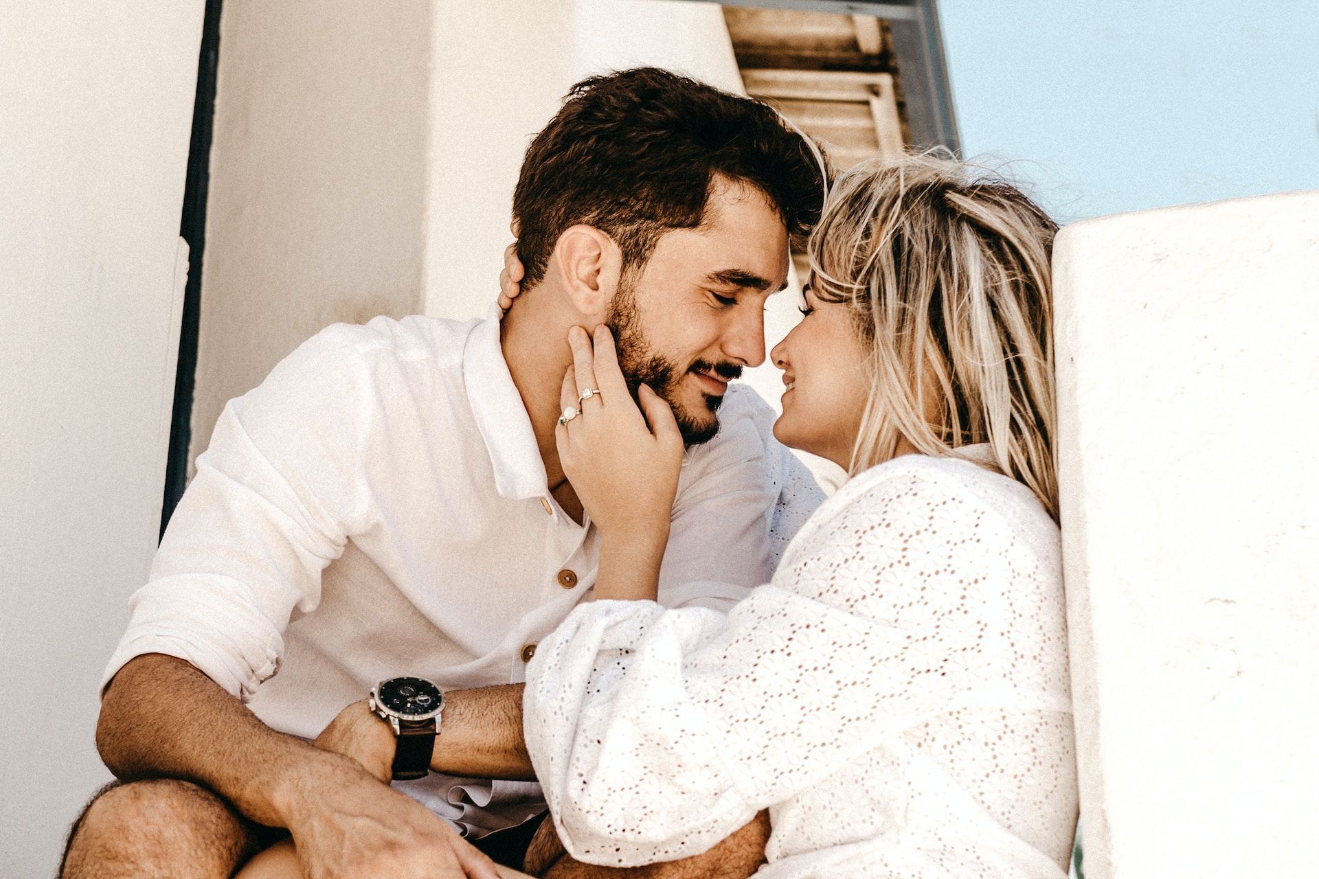 Couple gazes at each other, touching faces. They sit outdoors near a white wall, both wearing white clothing.