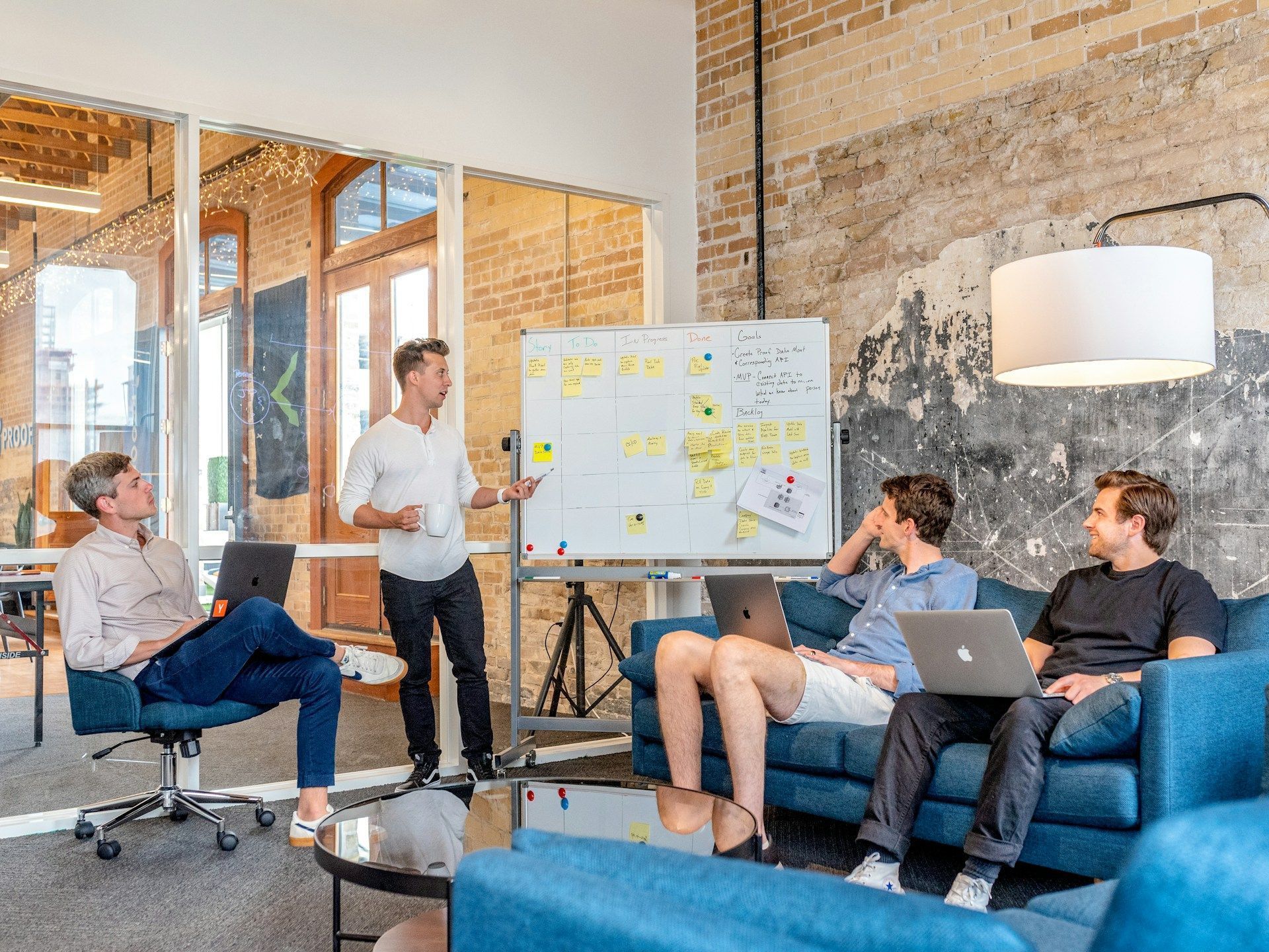 Four men in an office setting: one presenting at a whiteboard, three others seated with laptops, brick and glass walls.
