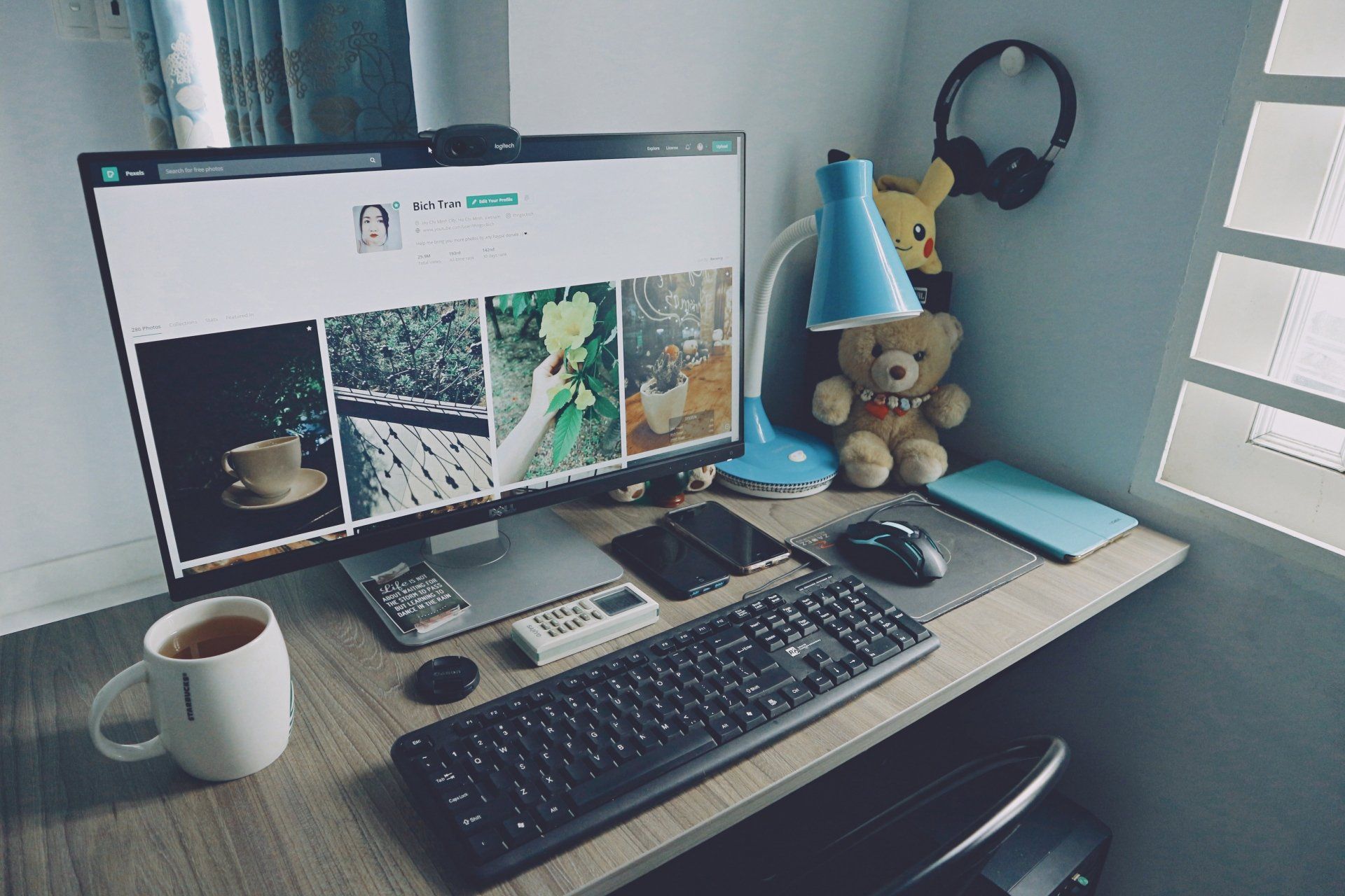 Desk with computer, lamp, teddy bear, headphones, and cup.