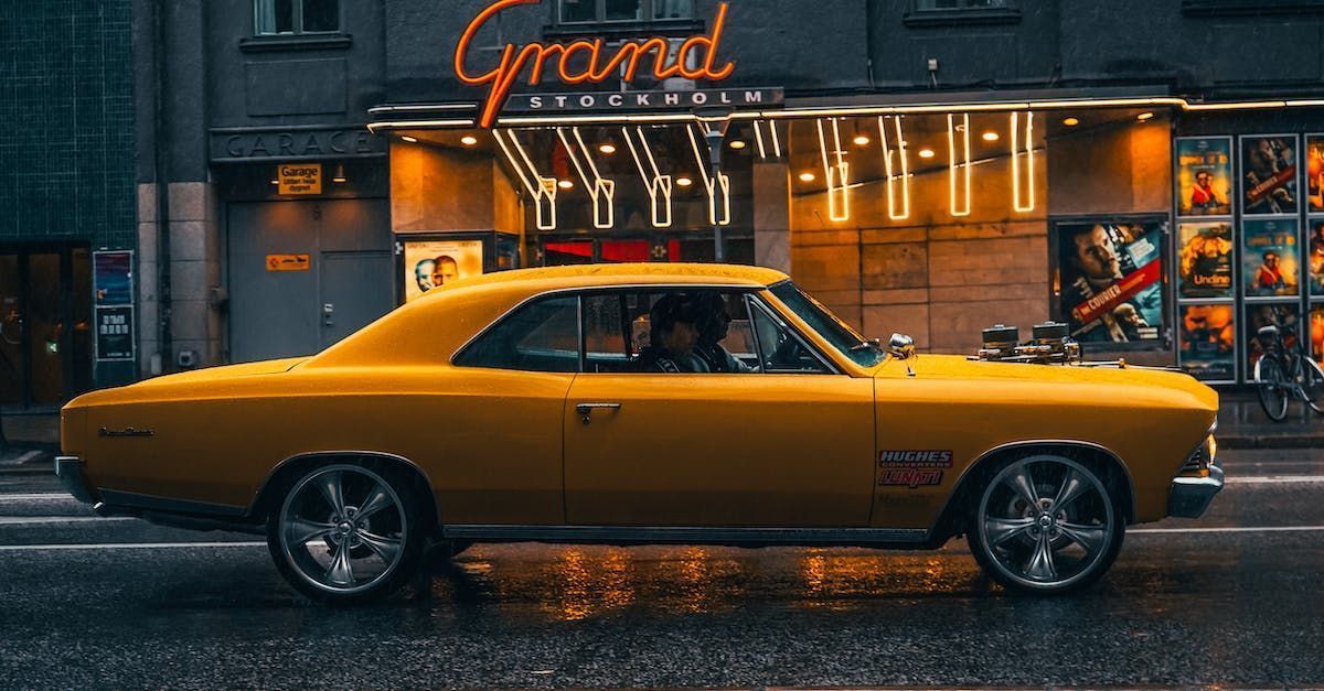 Yellow classic car driving past a theater with neon signs, wet street.