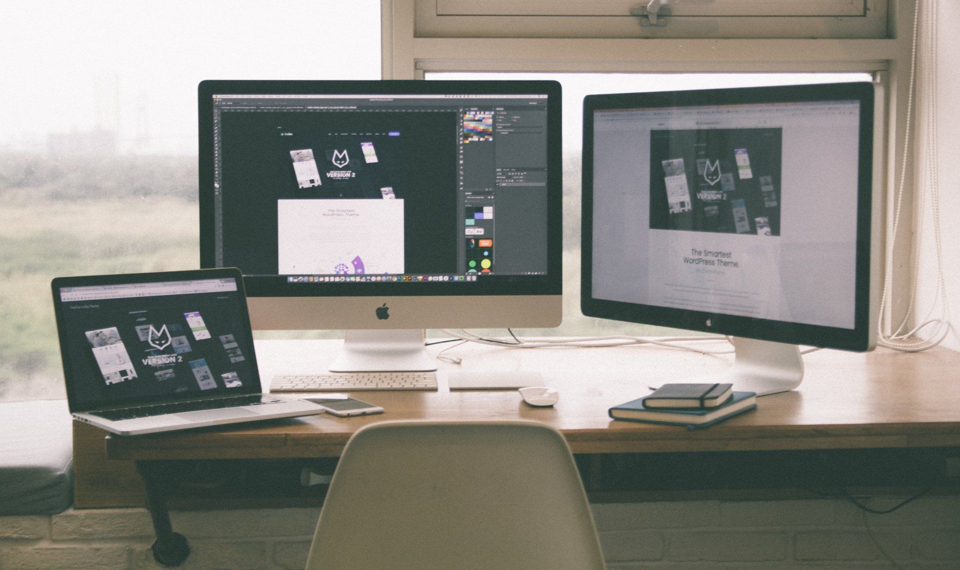 Desk with three screens displaying digital content, a laptop, and a notebook near a window.