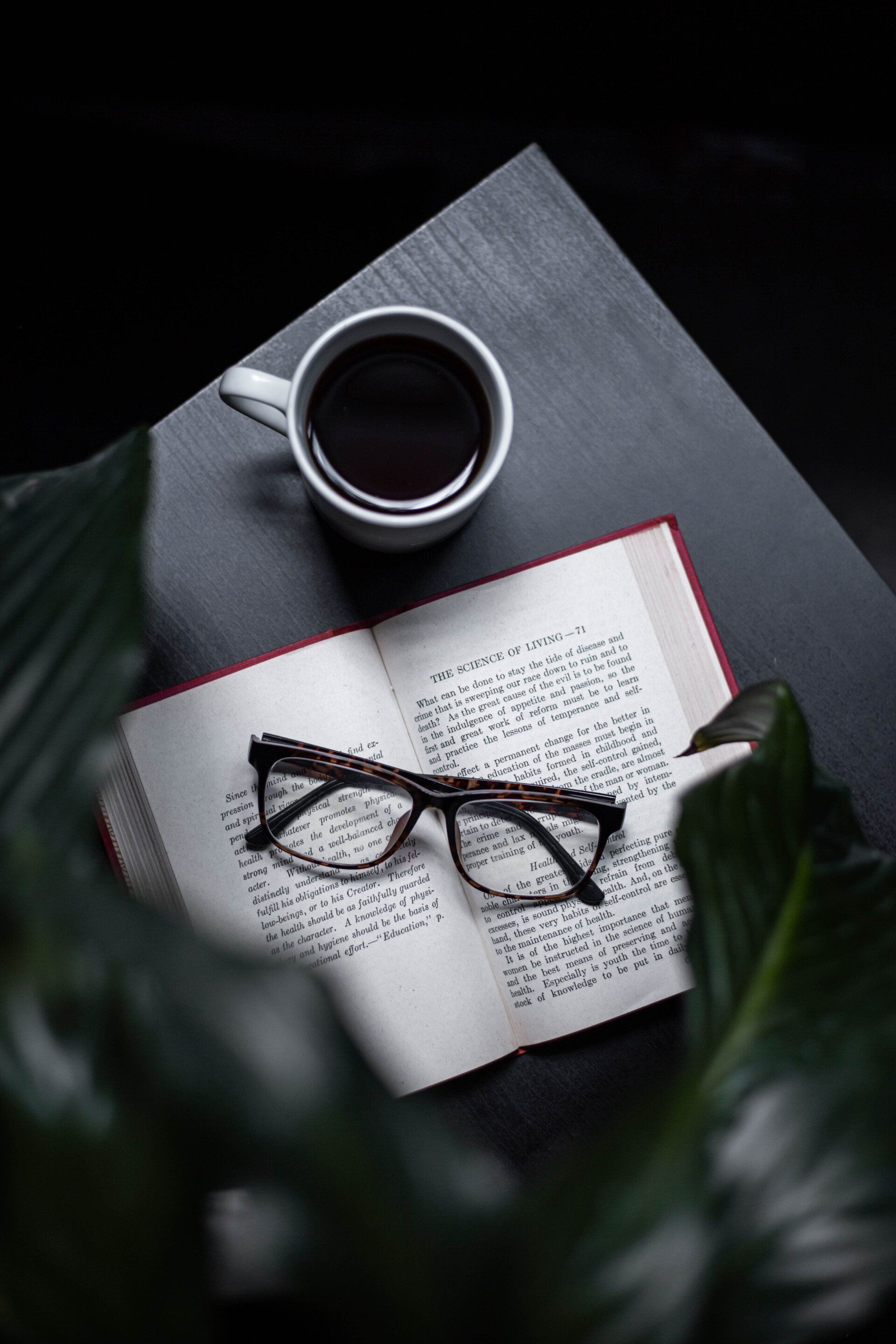 Coffee cup, open book with glasses, on a dark table, surrounded by green leaves.