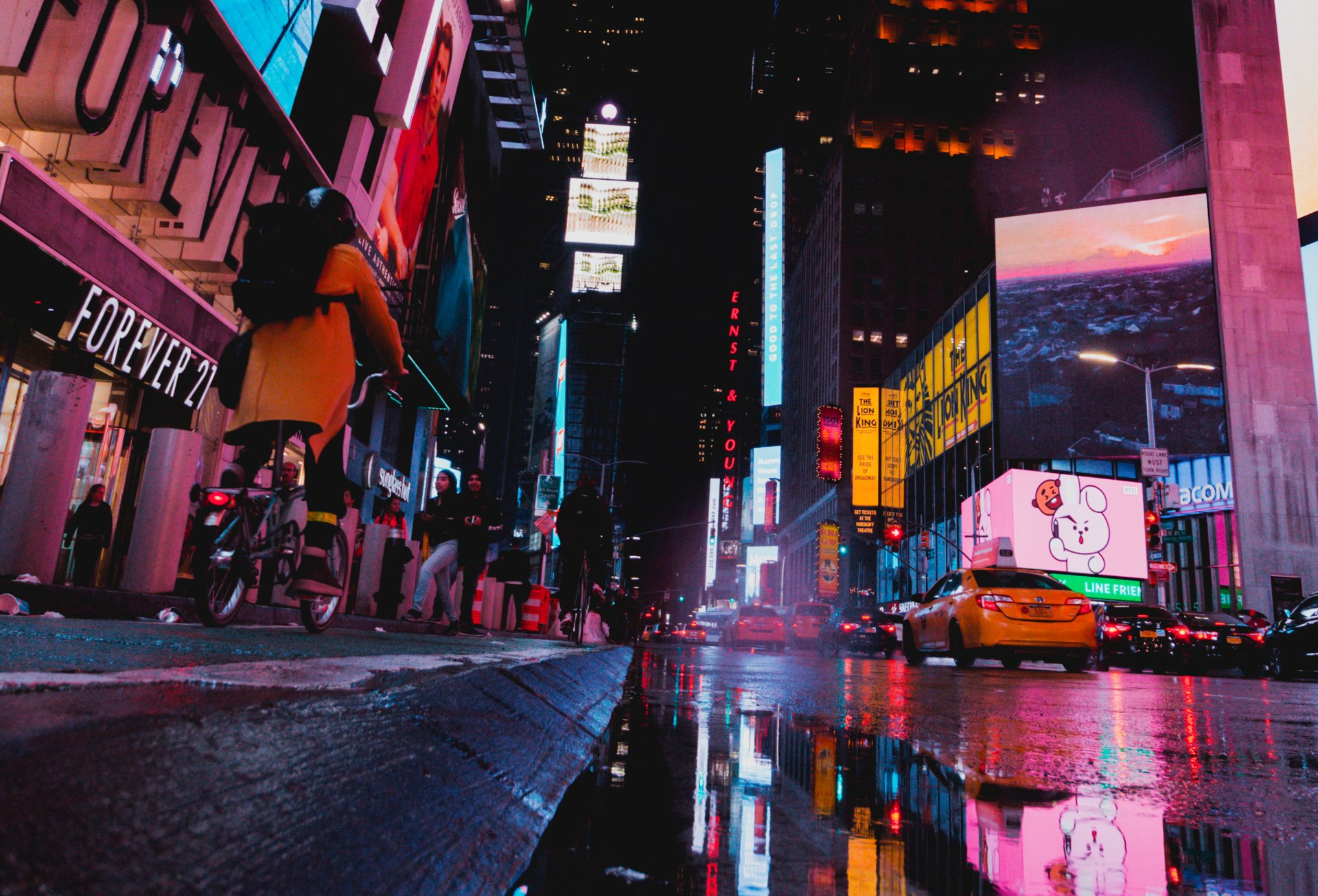 Times Square at night, with bright billboards and a yellow taxi on a wet street. A person rides a scooter.