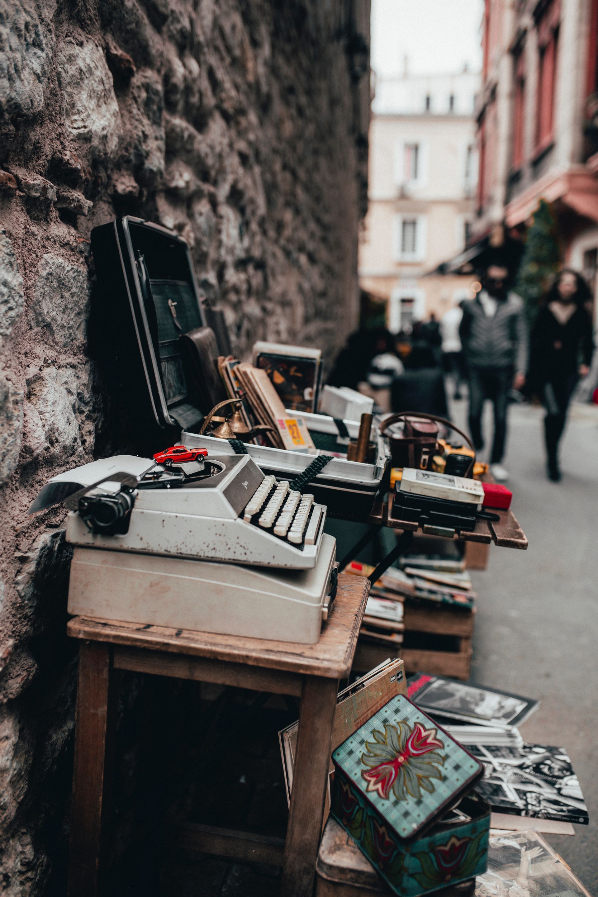 Antique typewriter and other items on a wooden stand in an alley; people walking in the background.