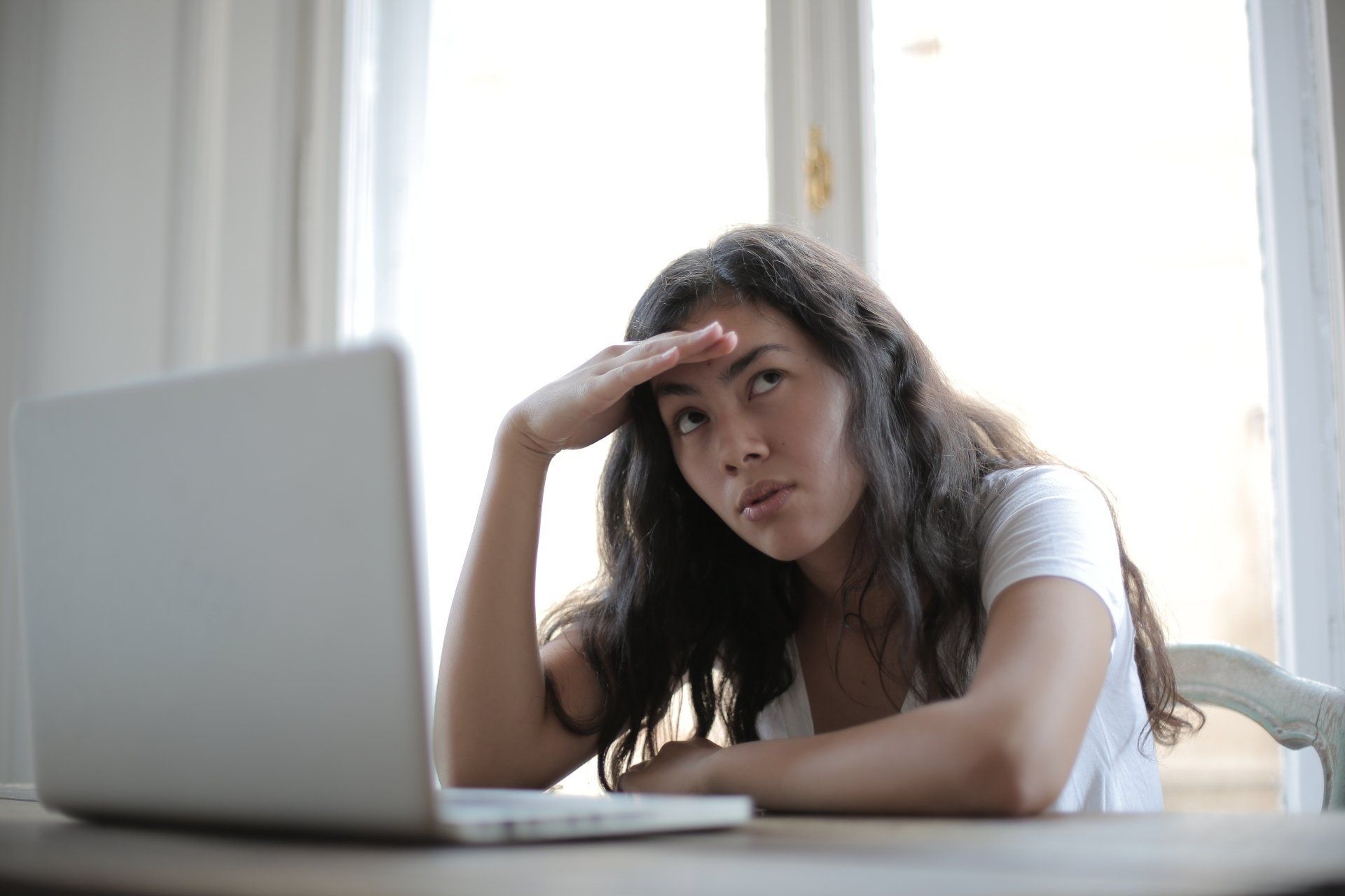 Woman at laptop, looking away with hand over her brow, appearing frustrated.
