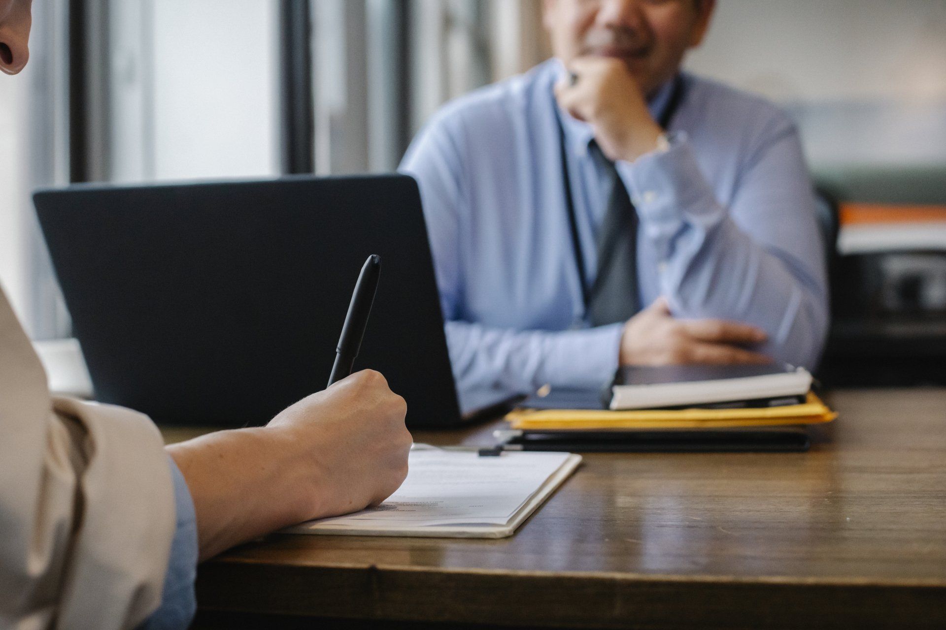 Person writing on a document across from another person at a desk with a laptop.