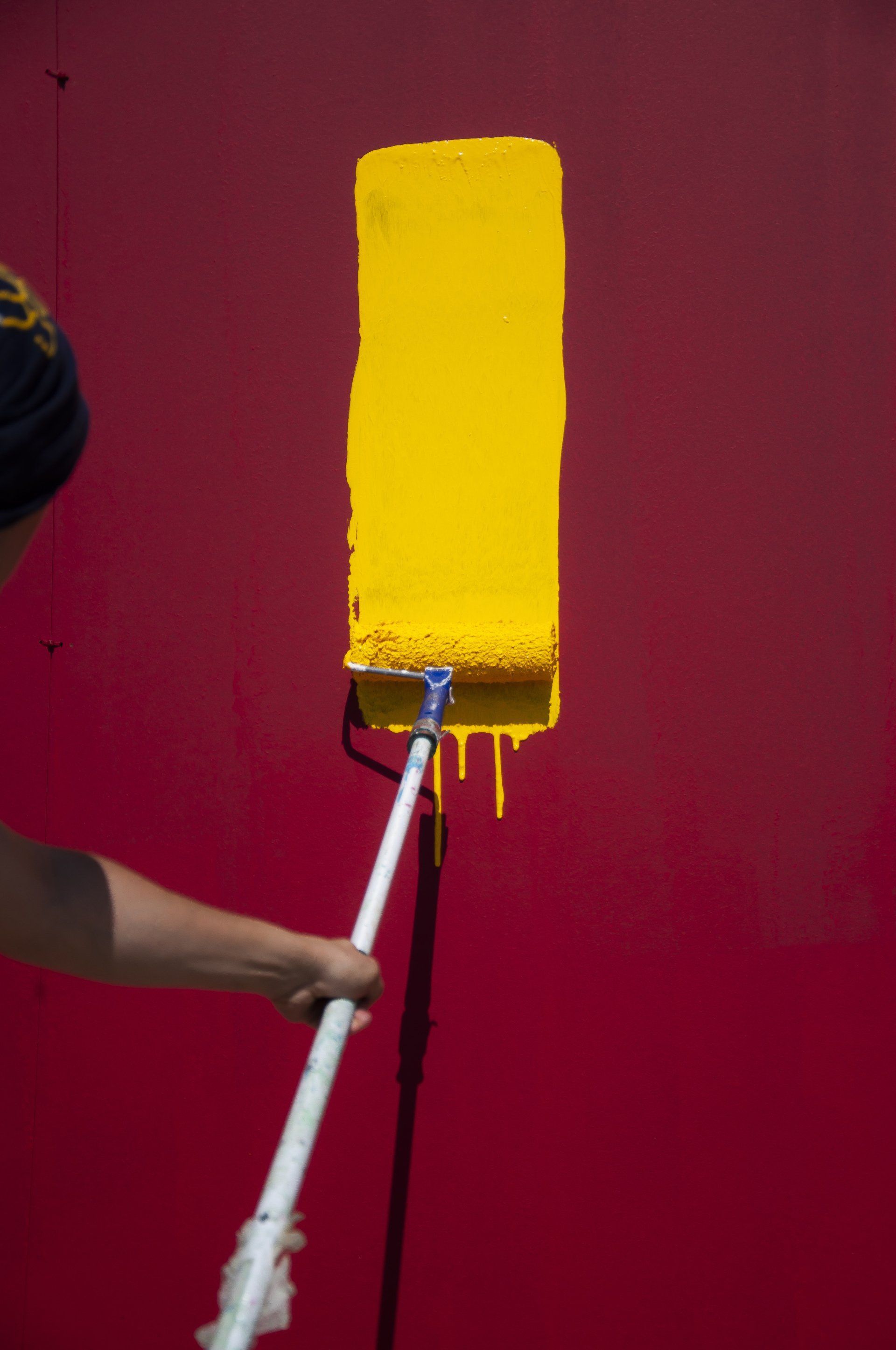 Person painting a red wall with a yellow paint roller; yellow paint drips.
