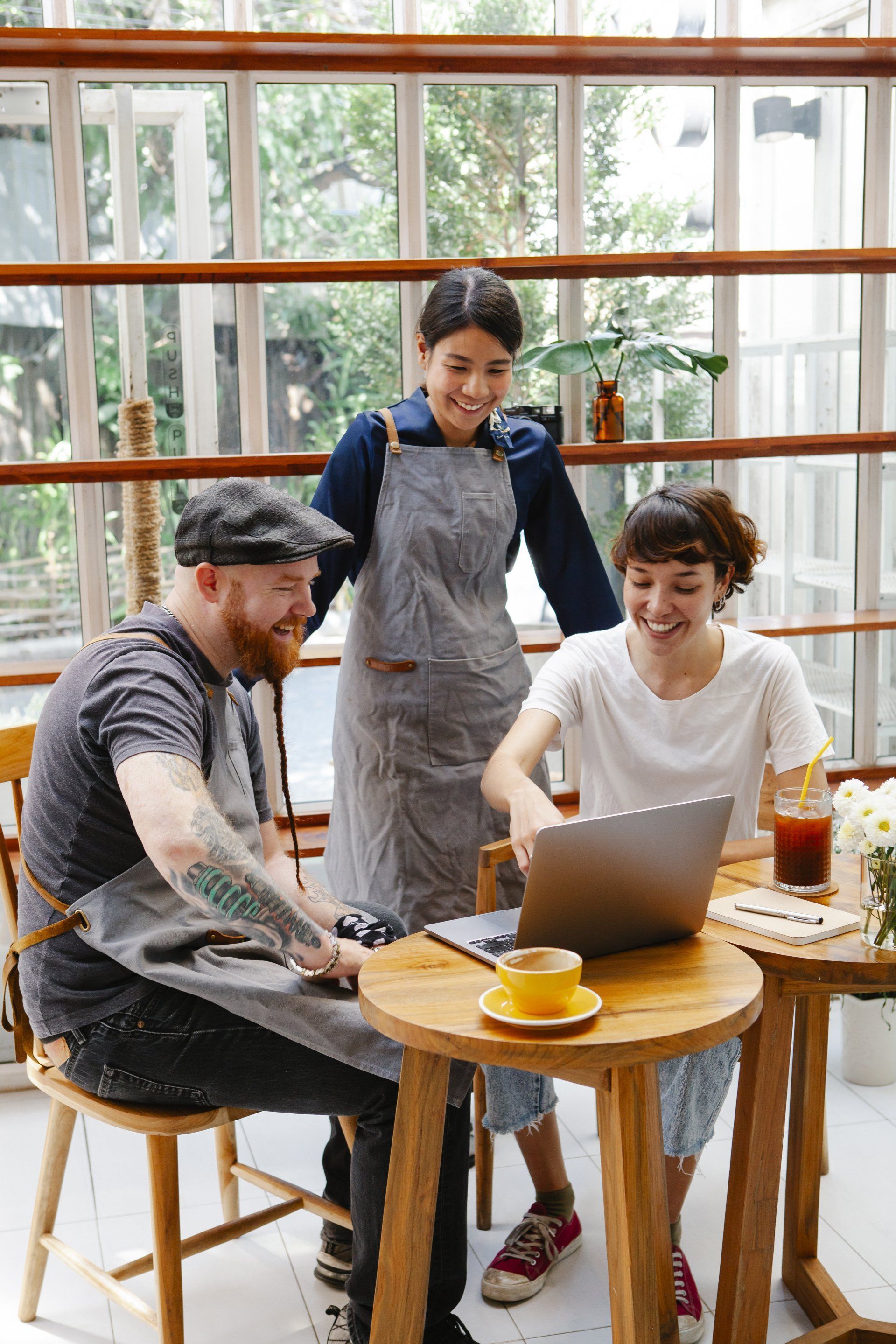 Three people in aprons look at a laptop in a cafe with a window background.