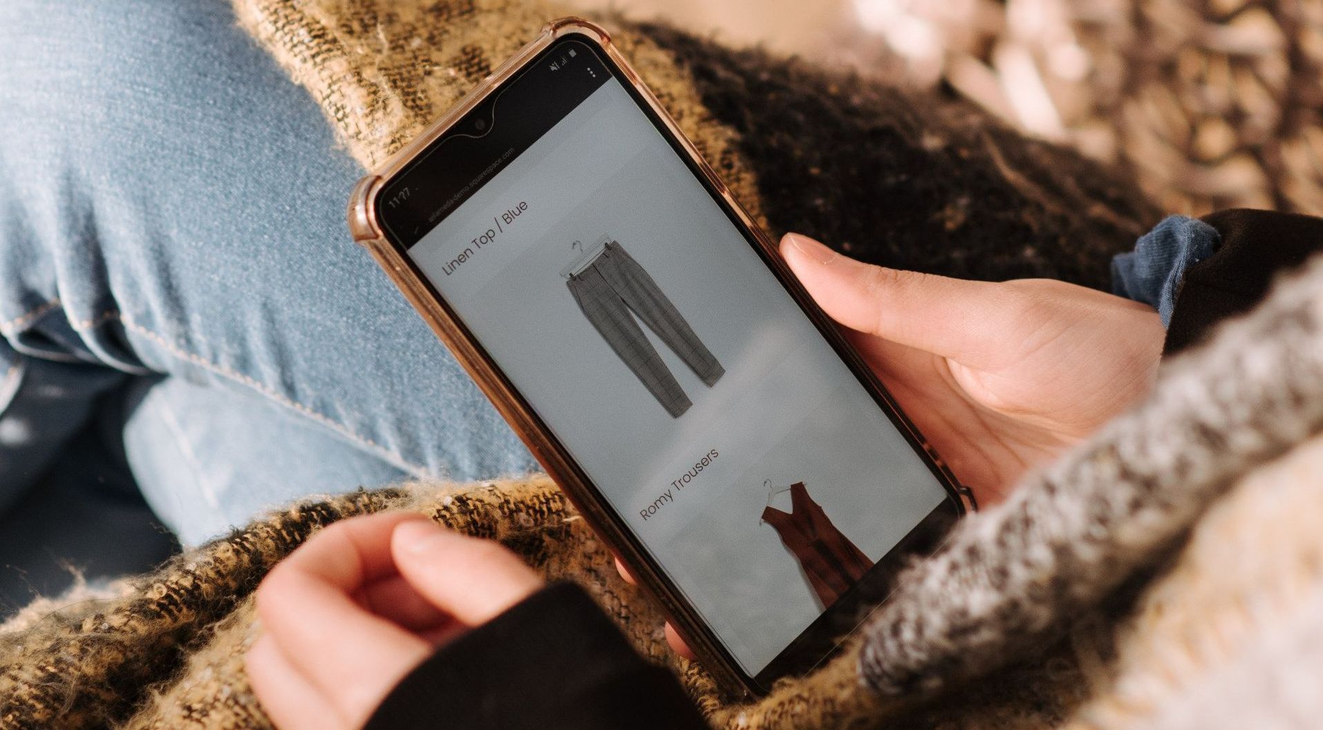 Person holding phone displaying clothing options. Sitting outdoors with cozy blanket, looking at pants.