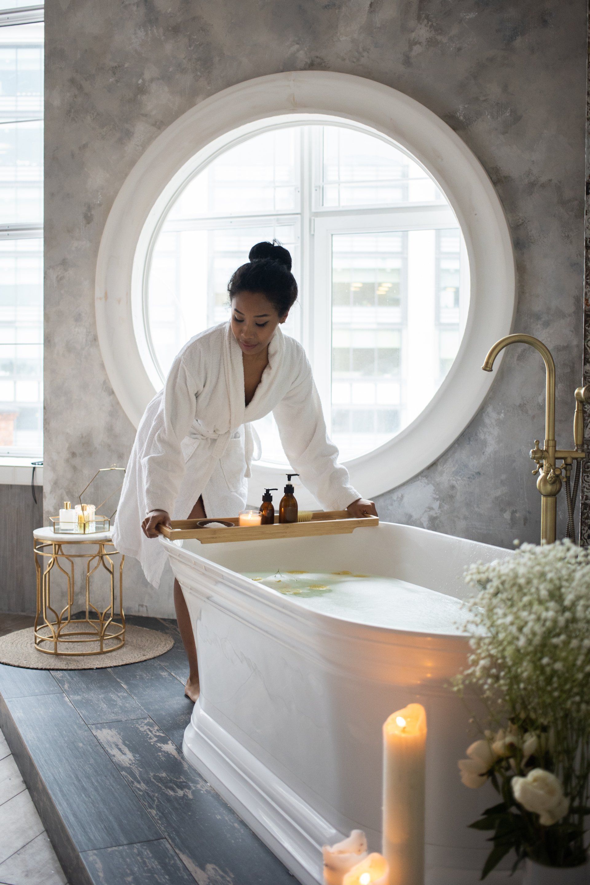 Woman in robe, setting up bath tray with lotions and candles near a tub.