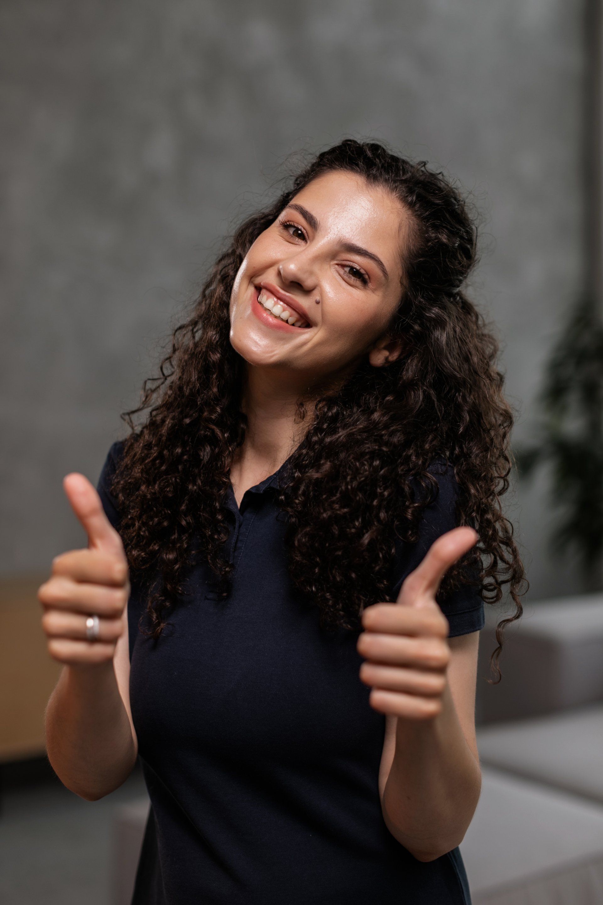 Woman with curly hair smiles, gives two thumbs up. Dark blue shirt, neutral backdrop.