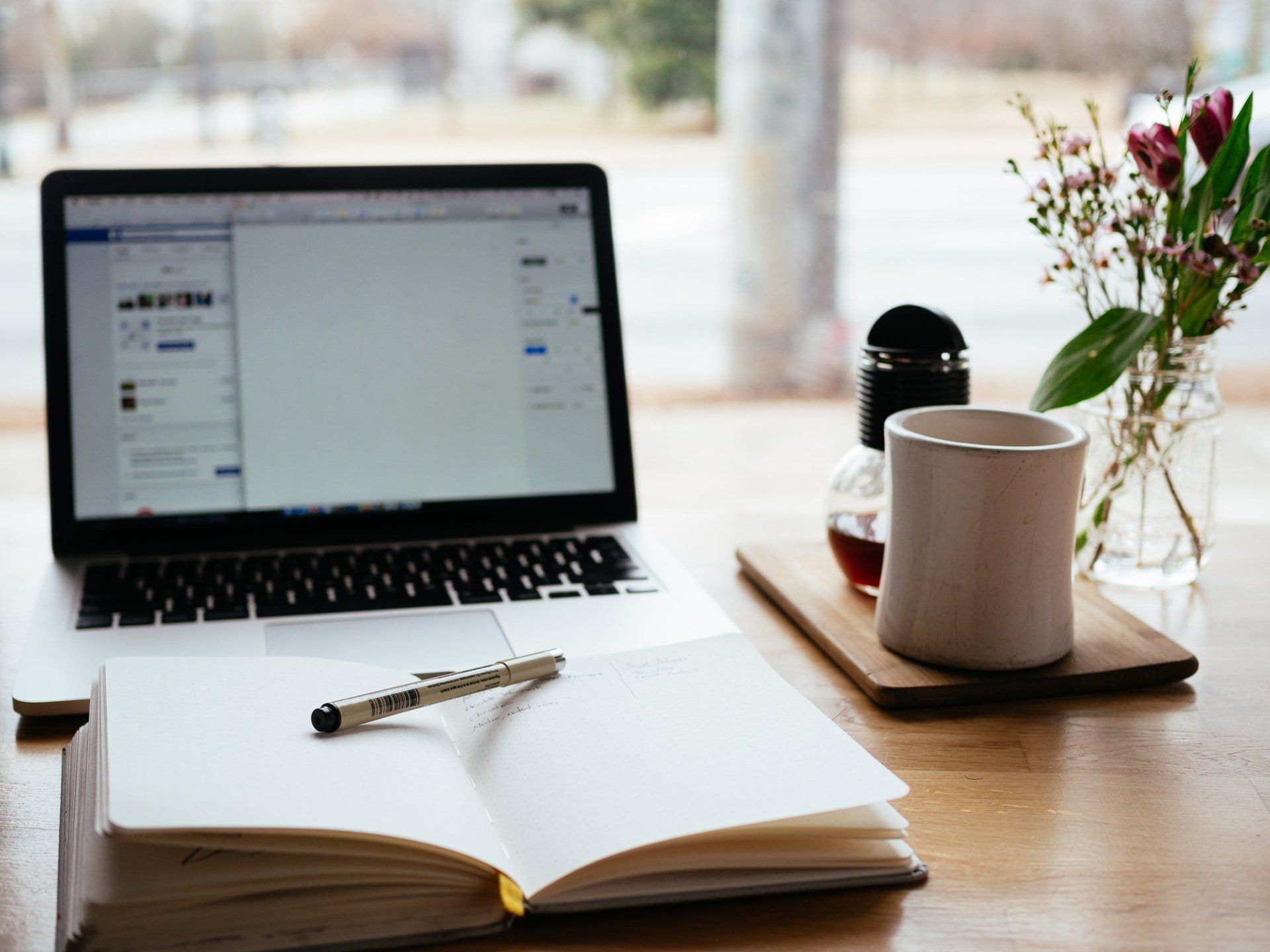 Laptop, open notebook, coffee cup, and flowers on a wooden table near a window.