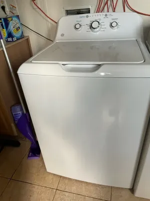 A white GE top-load washing machine sits on a tiled floor in a corner with a purple mop leaned against its side.