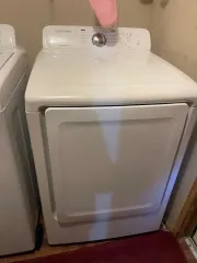 A white Samsung clothes dryer with a front-loading door and control panel on the top back, positioned in a laundry room.