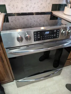 A stainless steel electric range with a glass cooktop and digital control panel, installed in a kitchen.