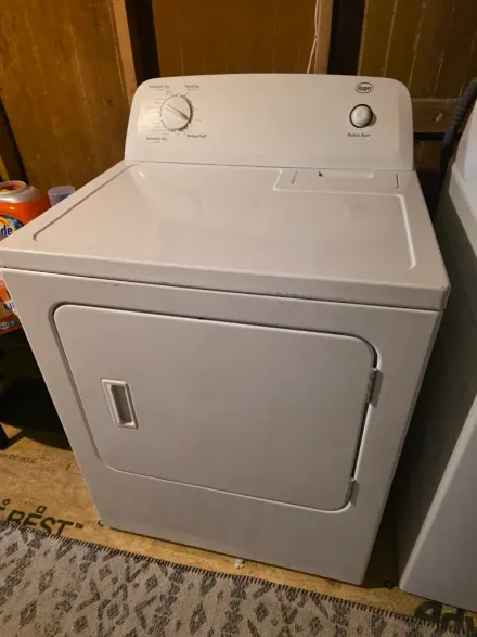 A white clothes dryer sits on a basement floor next to a washing machine, featuring a front-loading door and top dials.