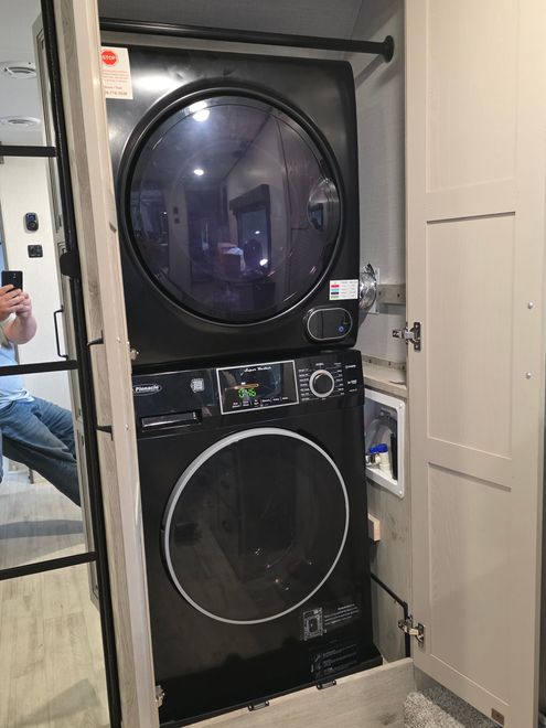 A stacked black washer and dryer unit installed in a closet within an RV, with a person reflected in a mirror.