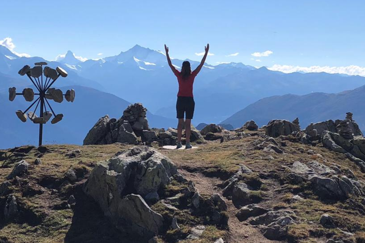 Een persoon staat met opgeheven armen op een rotsachtige bergtop vande Bettmerhorn  de Matterhorn en besneeuwde bergtoppen.