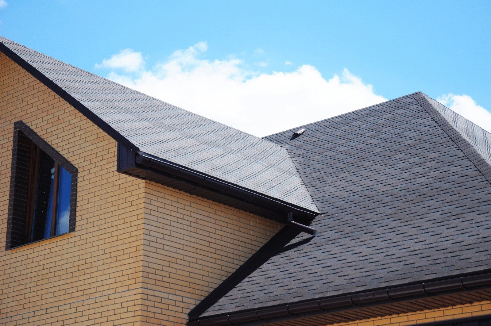 Tan brick house with dark gray shingled roof and a blue sky background.