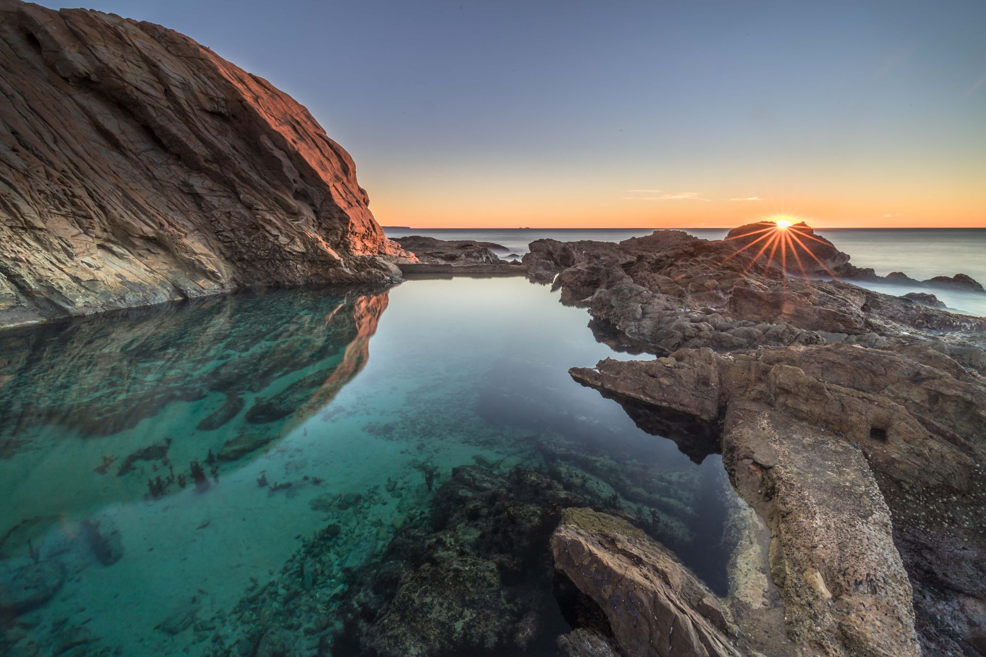 Rocky coastline with a turquoise tide pool reflecting the setting sun's golden rays.