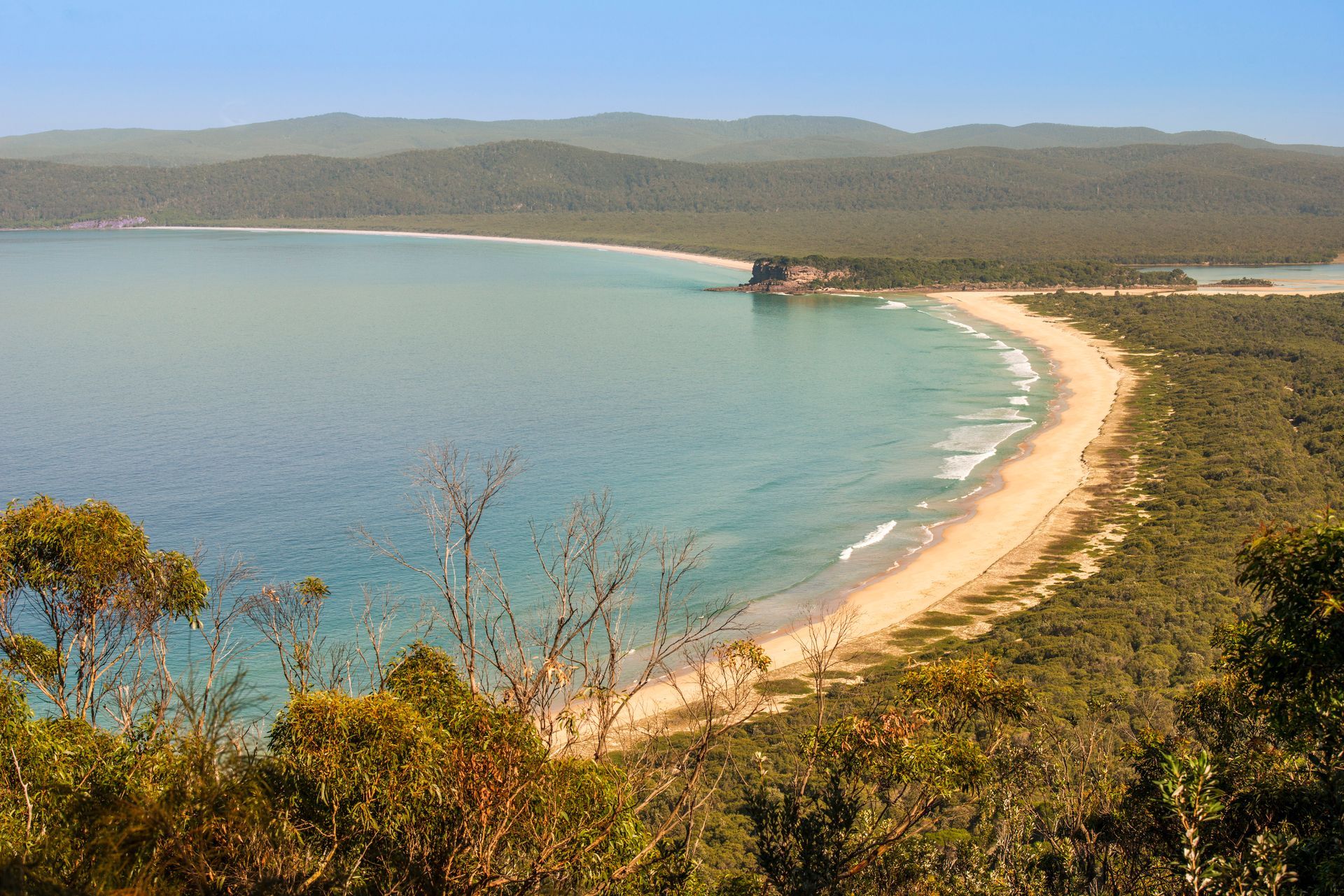 Curved sandy beach, turquoise water, and forested shoreline under a blue sky.