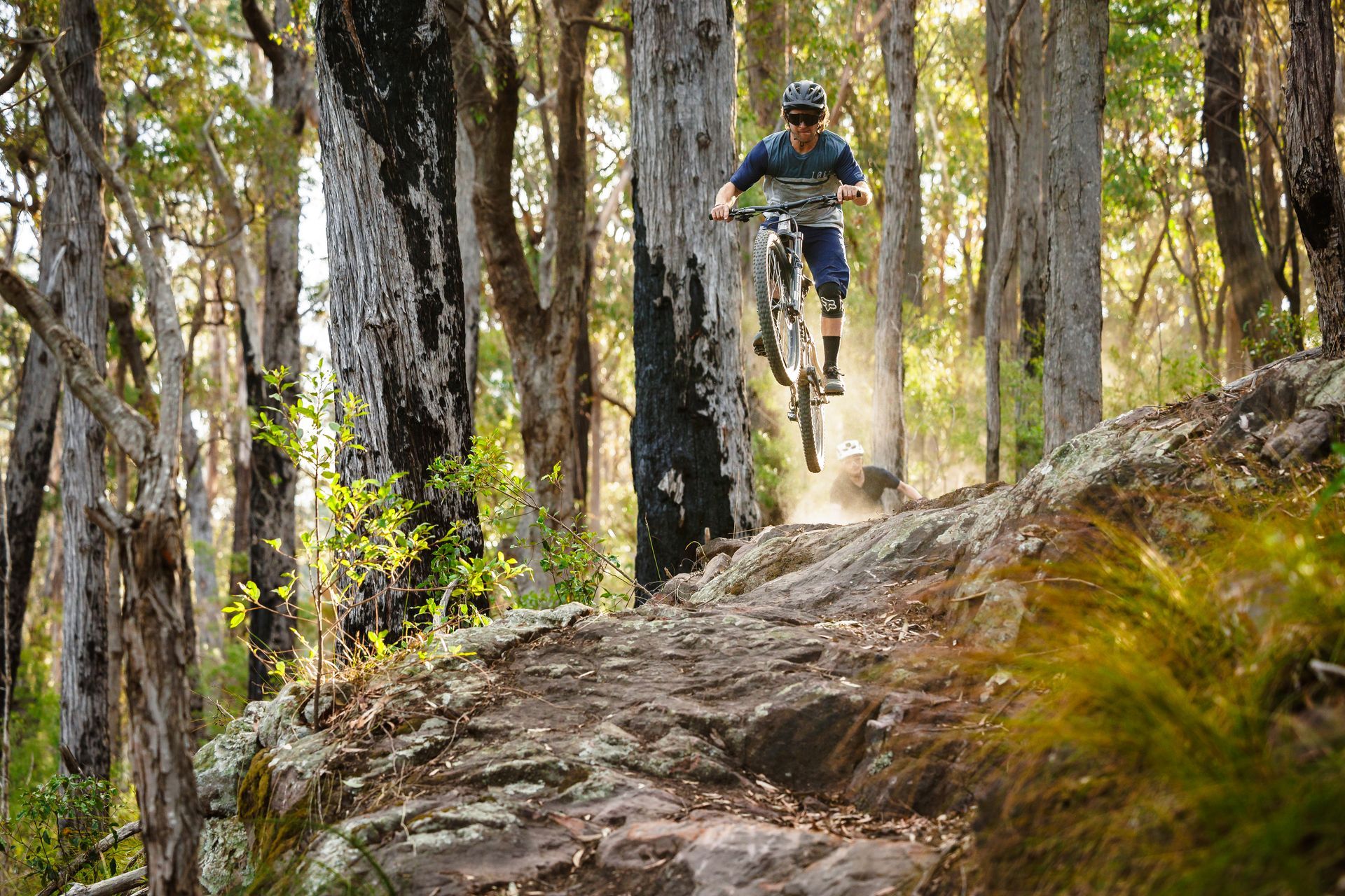 Mountain biker airborne over rocky terrain in a forest.