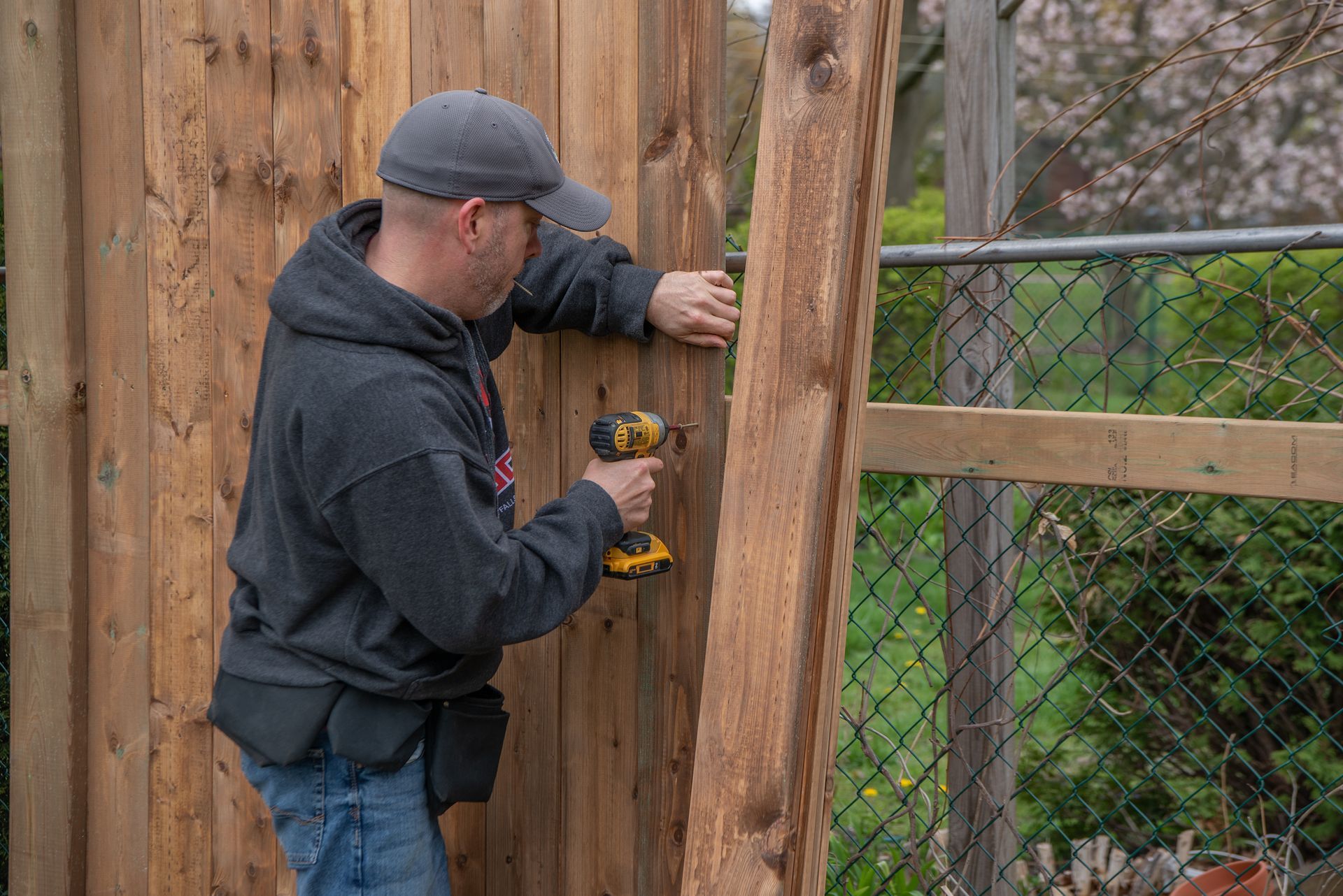 Man drills wood plank onto fence outdoors, wearing cap and tool belt.