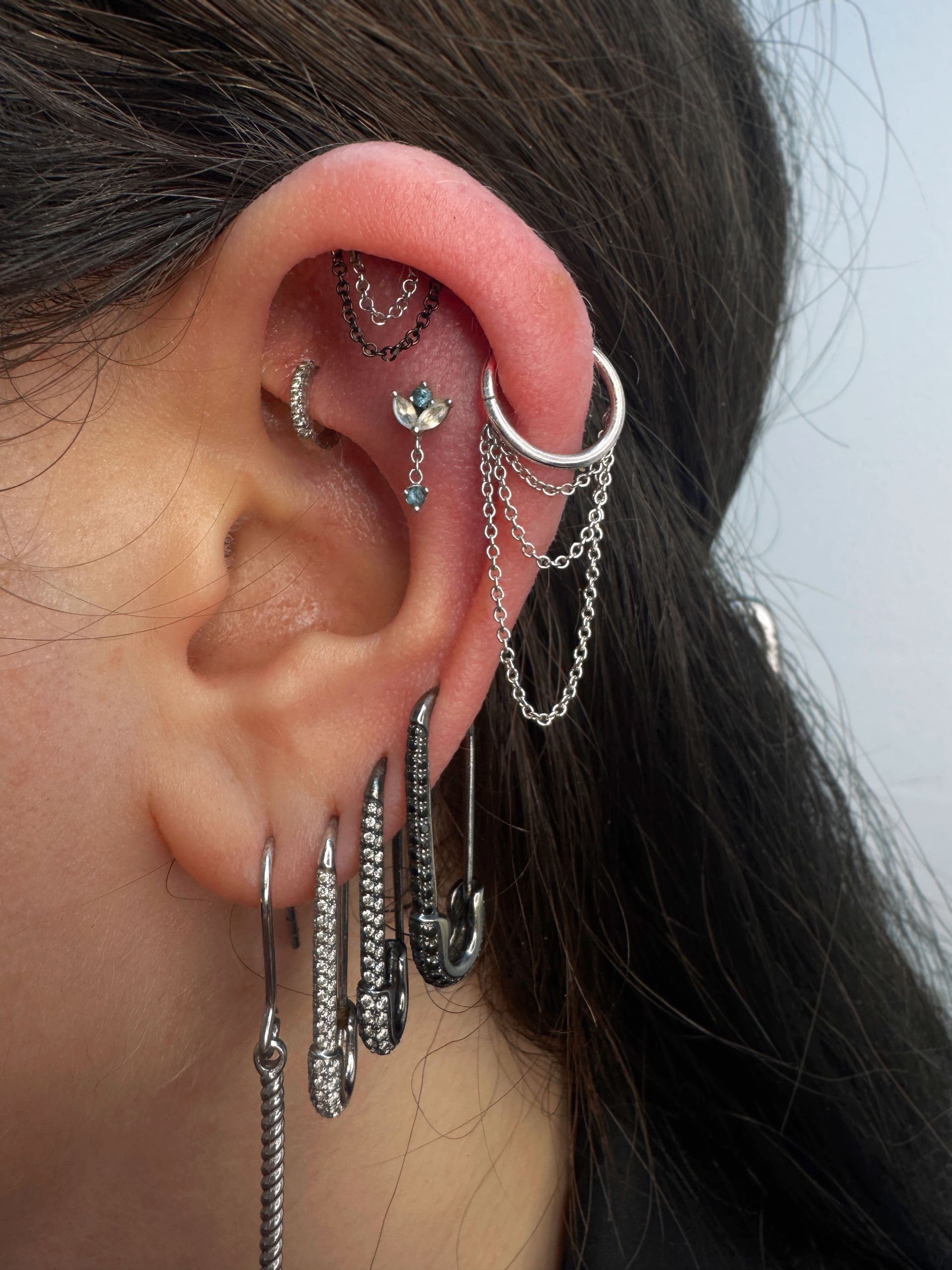 Close-up of a person's ear with multiple silver earrings, including chains, hoops, and safety pins.