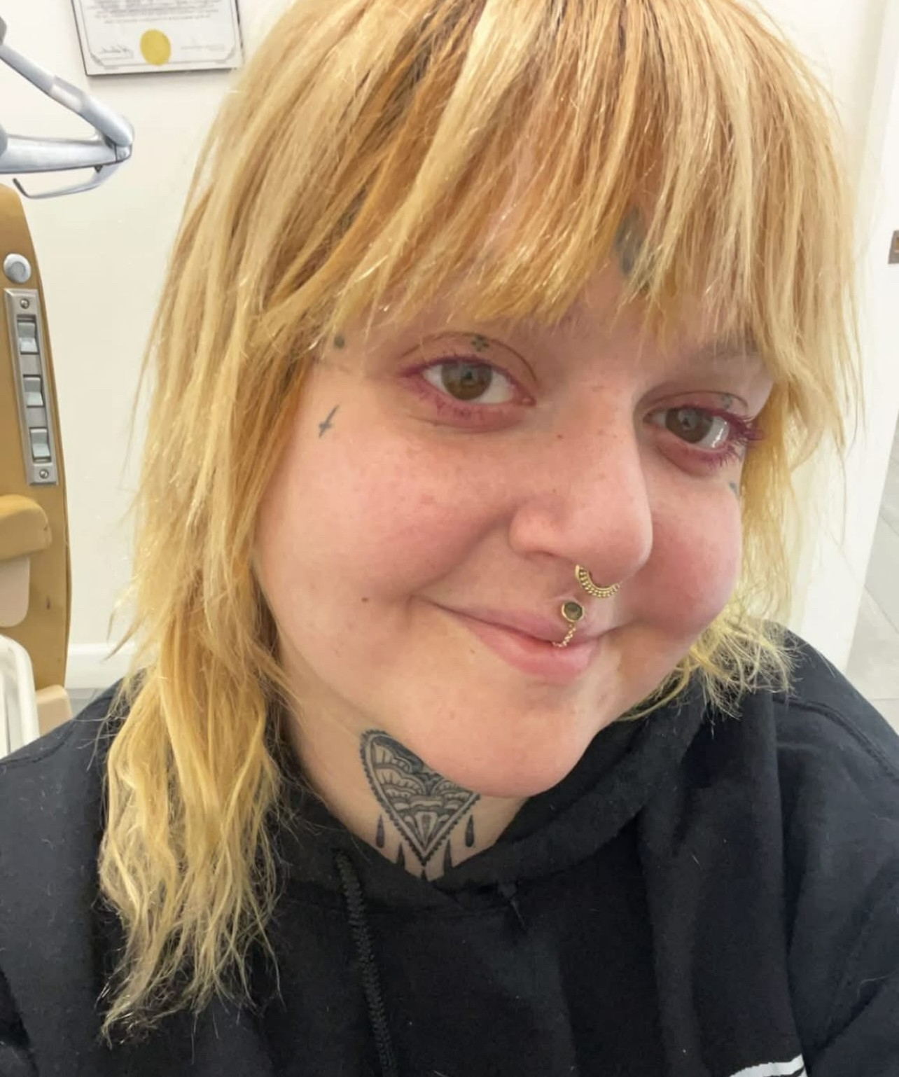 Person with blonde hair and facial piercings, smiling softly in a black hoodie indoors.