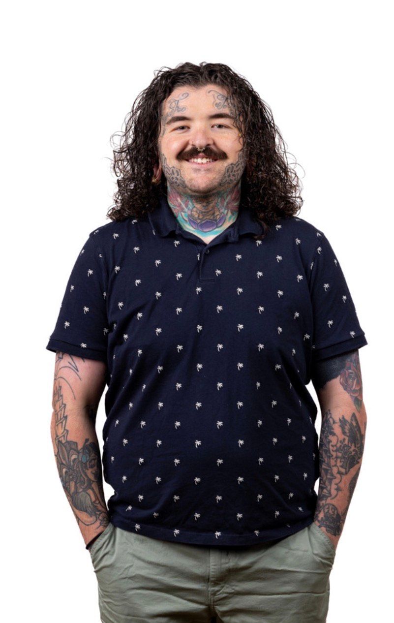 Man with curly hair and tattoos smiles, hands in pockets, wearing a navy polo and khaki pants.