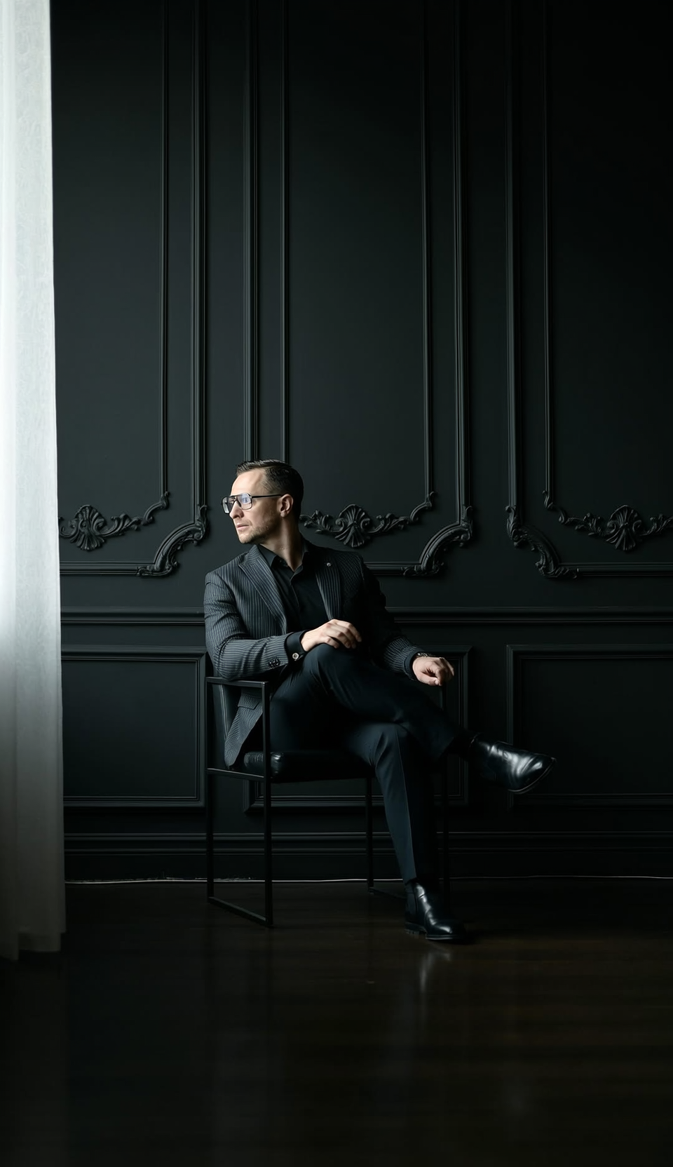 Man in a dark suit sits in a chair near a window, looking to his right. Dark walls.