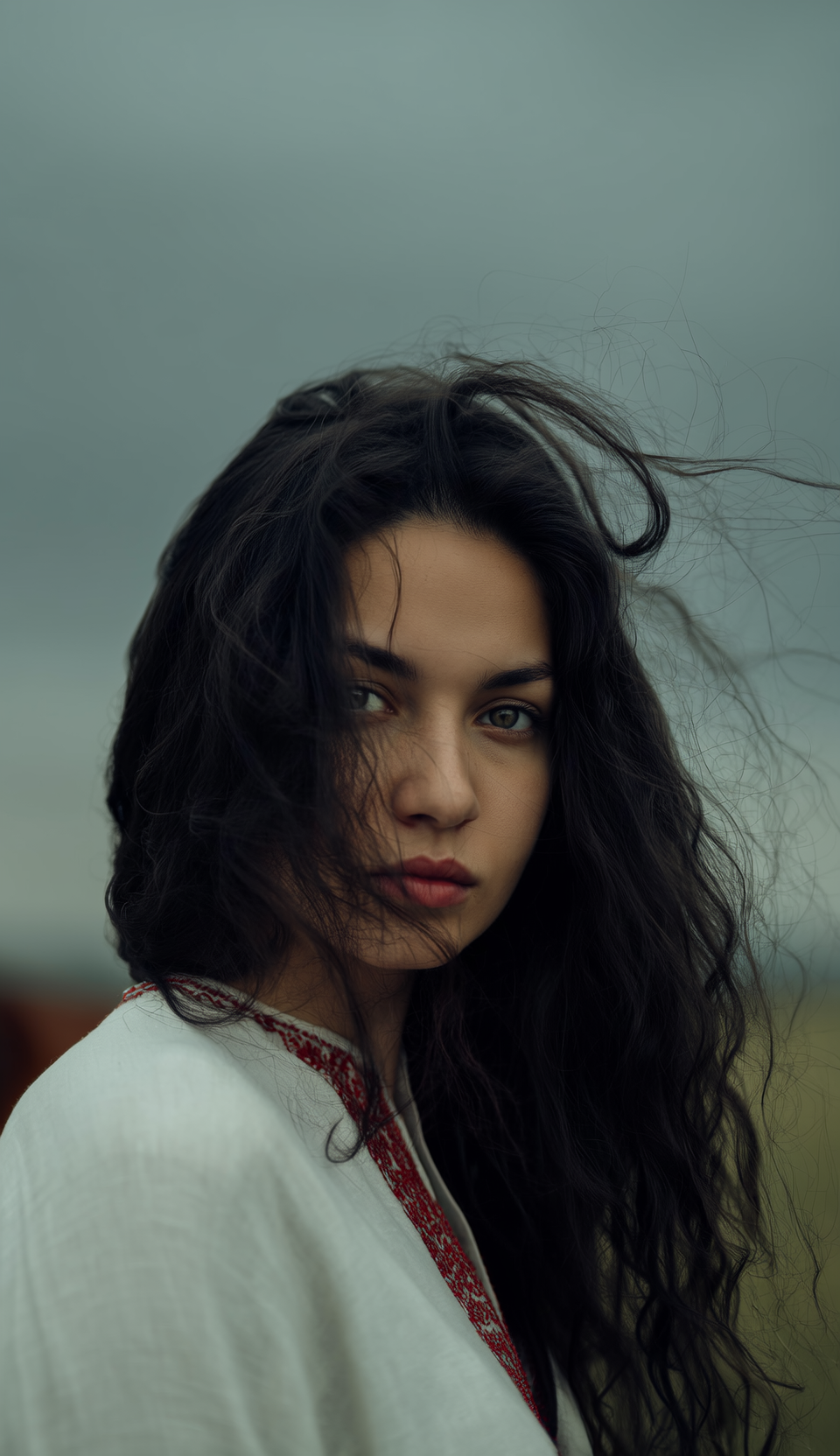 Woman with dark hair looking at camera, wearing white embroidered garment outdoors against overcast sky.