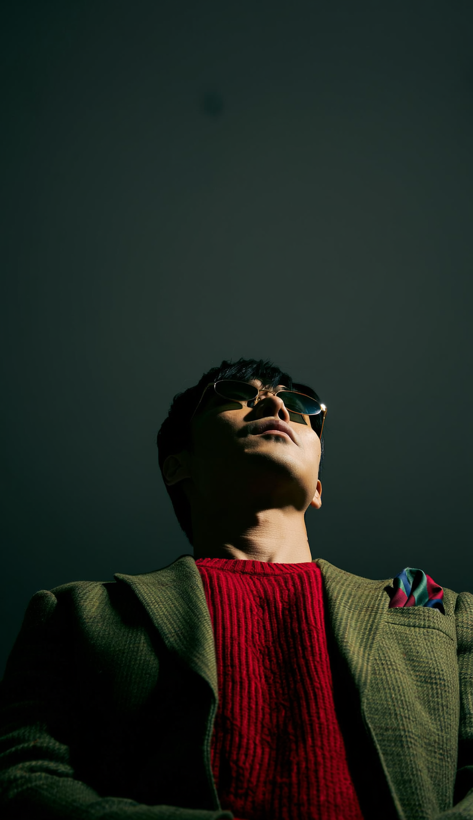 Man looking upwards, wearing sunglasses, red sweater, tweed jacket, and pocket square.