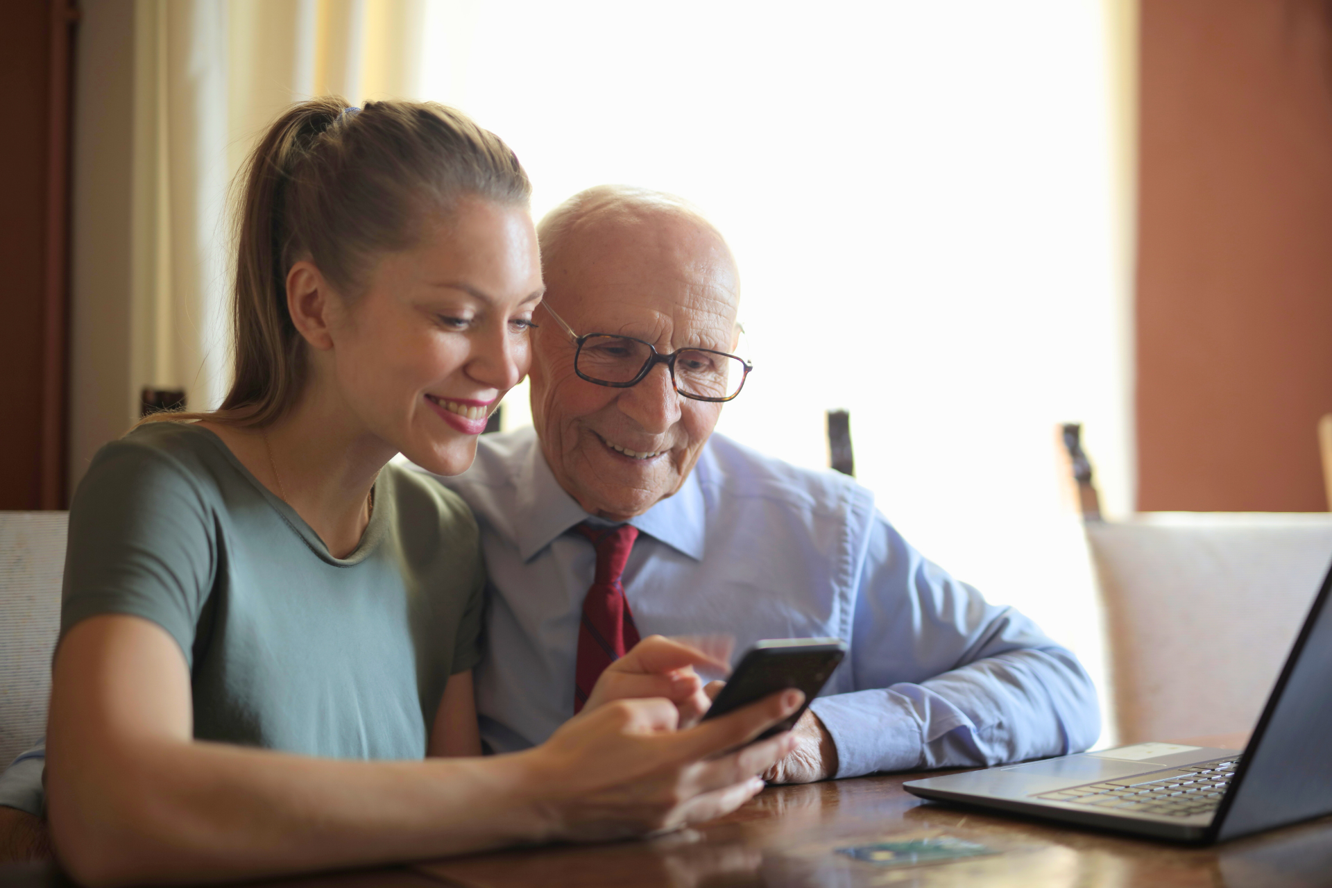Junge Frau zeigt einem älteren Mann ein Smartphone; beide lächeln und sitzen mit einem Laptop an einem Tisch.