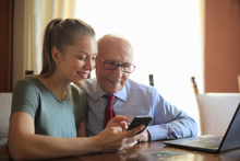 Junge Frau zeigt einem älteren Mann ein Smartphone; beide lächeln und sitzen mit einem Laptop an einem Tisch.
