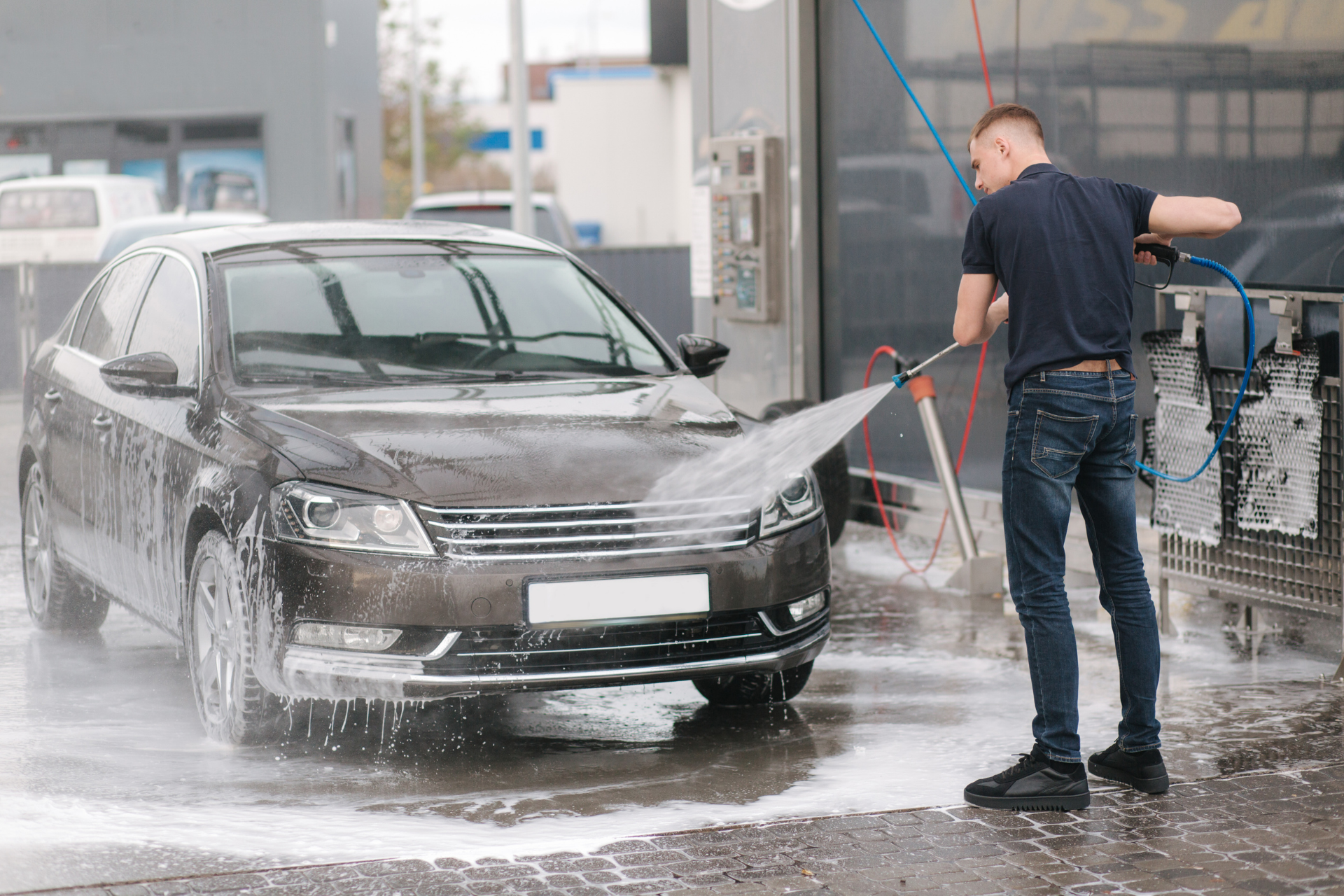 Mann wäscht in einer Autowaschanlage ein braunes Auto mit einem Hochdruckschlauch.